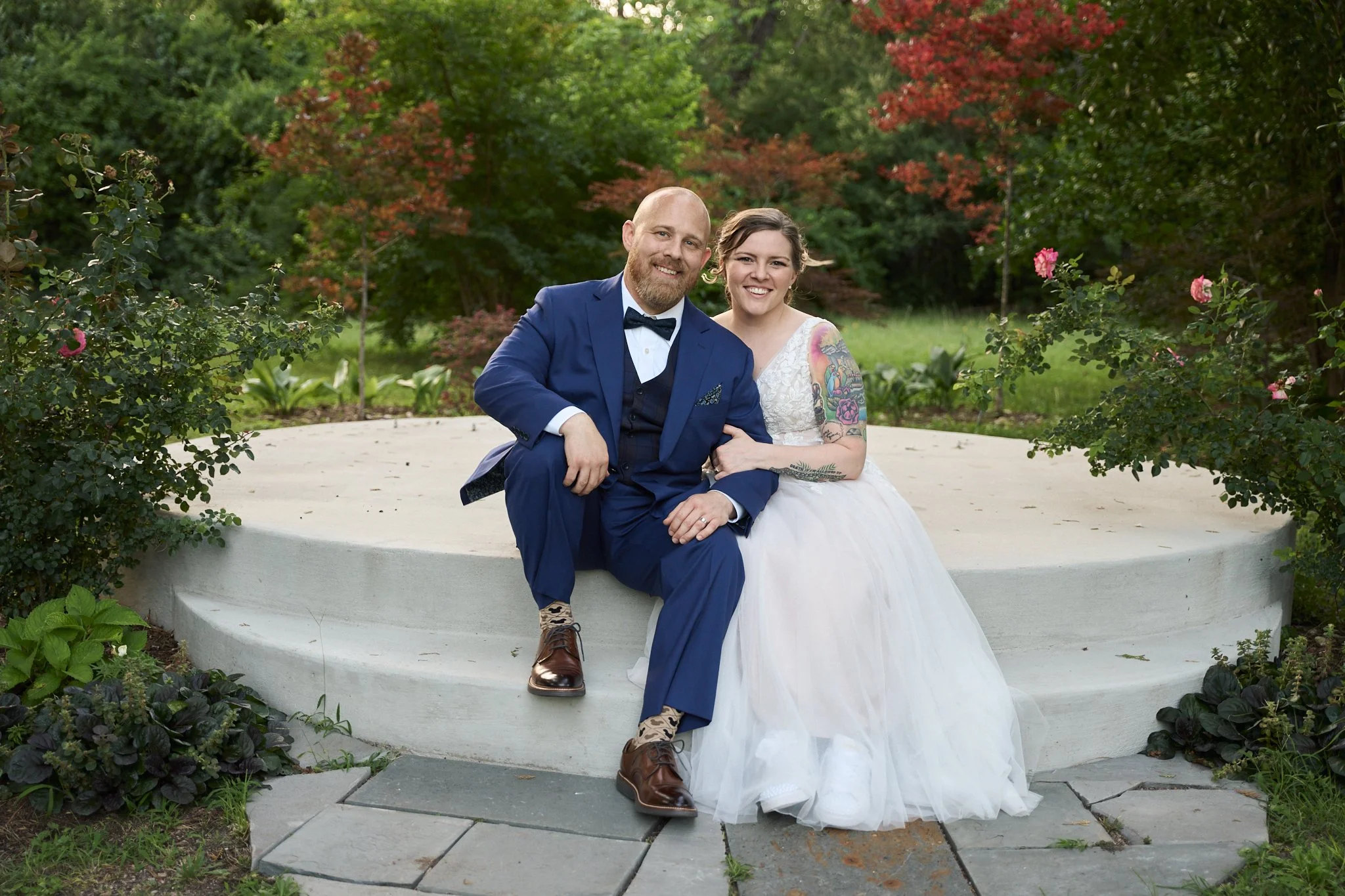 A couple dressed in wedding attire sitting on a curved white bench in a lush garden with green and red foliage.