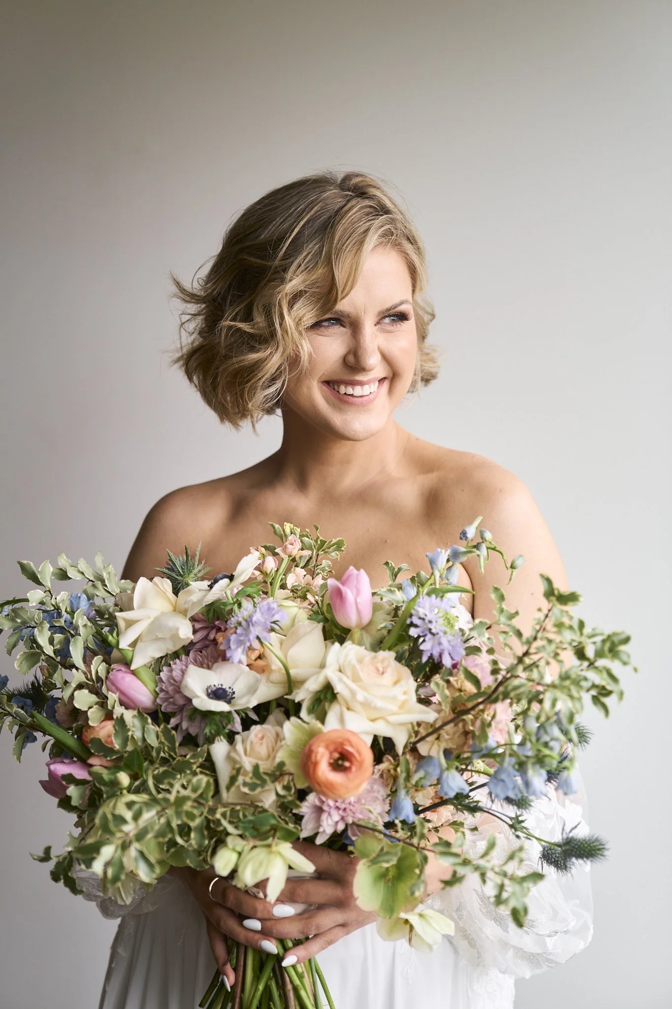 A woman with short wavy blonde hair smiling, holding a large bouquet of colorful flowers against a plain wall.