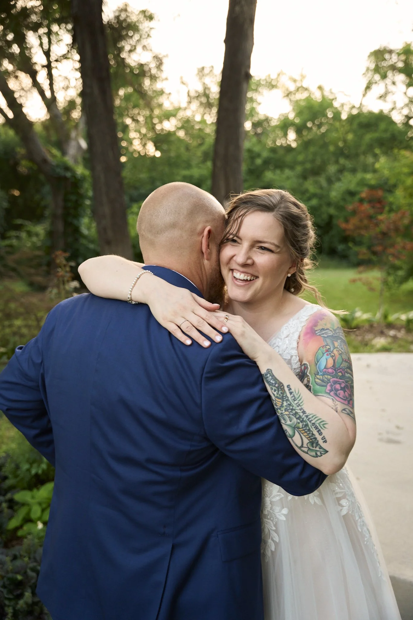 A joyful couple embracing outdoors with trees in the background; the woman has tattoos on her arms and wears a white dress, the man is dressed in a blue suit.