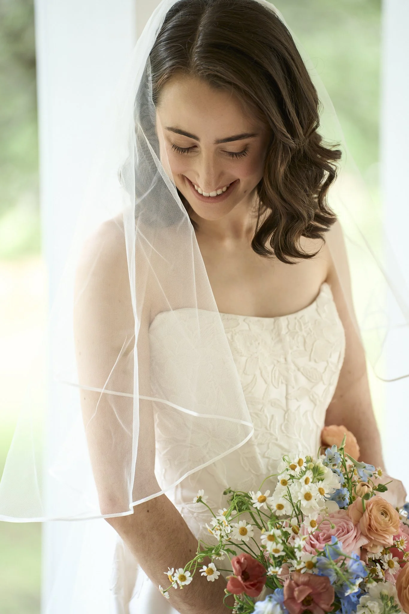 A smiling bride with wavy brown hair wearing a white lace strapless wedding dress and a sheer white veil, holding a bouquet of mixed flowers including daisies, roses, and baby’s breath.