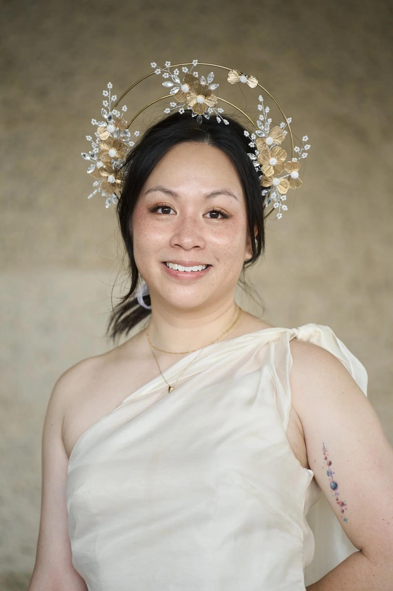 A woman with dark hair wearing a cream-colored satin dress, a gold heart-shaped necklace, and a decorative golden floral headpiece. She has a tattoo sleeve on her left arm and is smiling at the camera against a neutral background.
