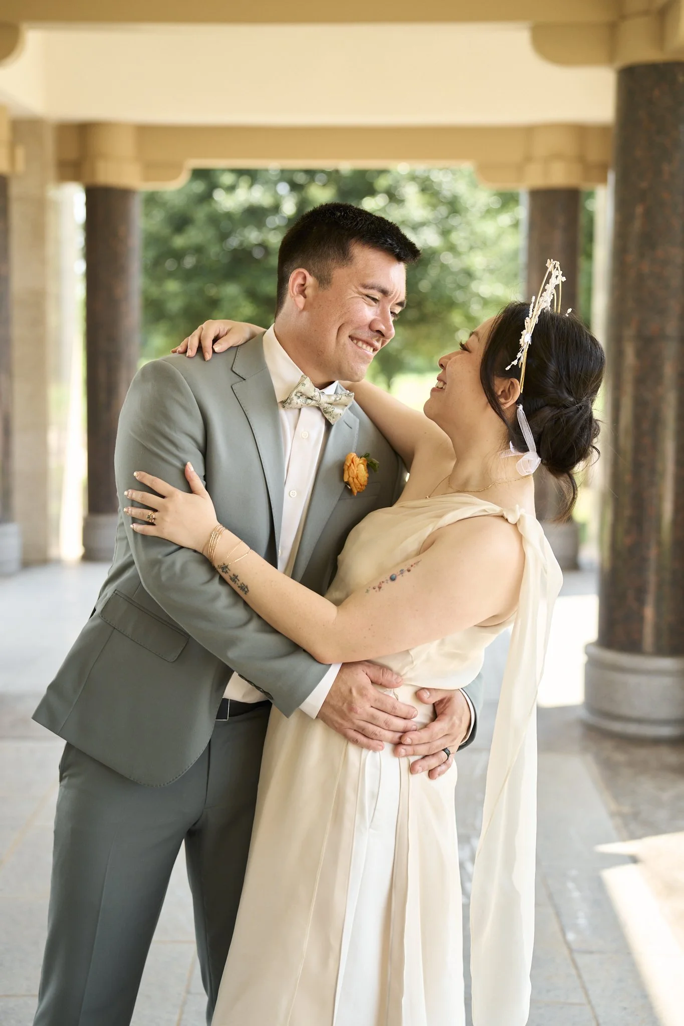 A couple dressed in wedding attire sharing a dance and smiling at each other outdoors with trees and columns in the background.