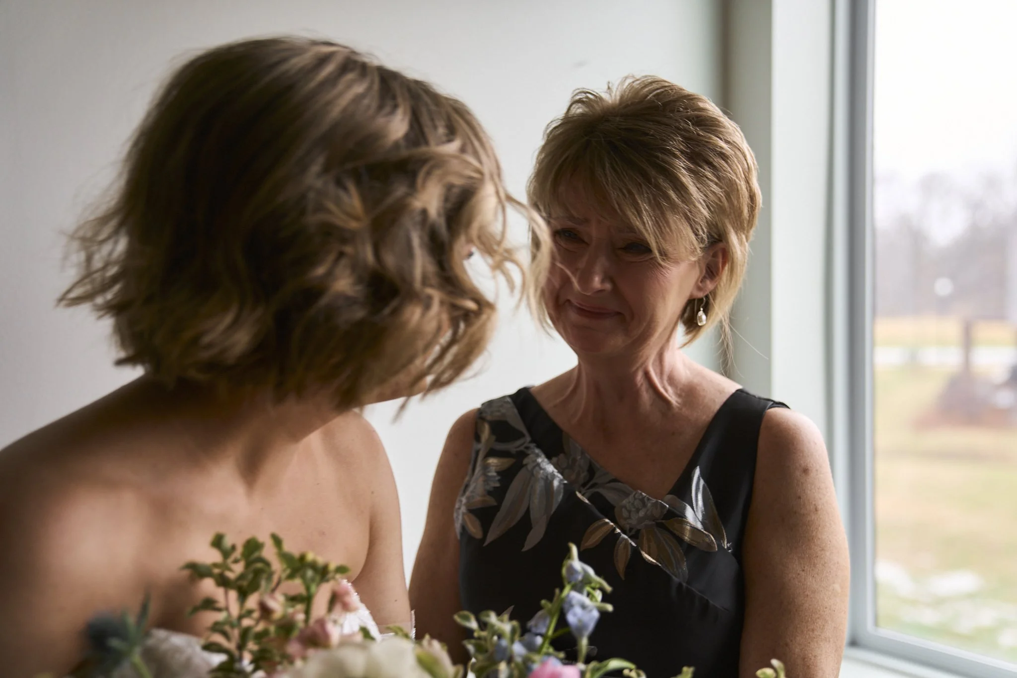 Two women, one older and one younger, are standing close together near a window, sharing an emotional moment. The older woman is smiling through tears, while the younger woman has her back to the camera, and they appear to be exchanging a meaningful 