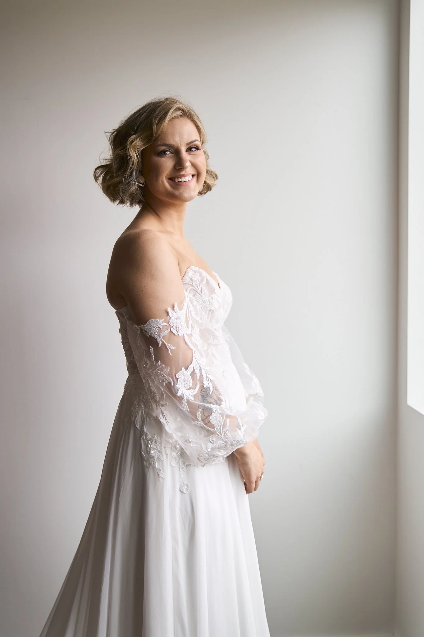 A woman in a wedding dress standing near a window with light streaming in, smiling at the camera.