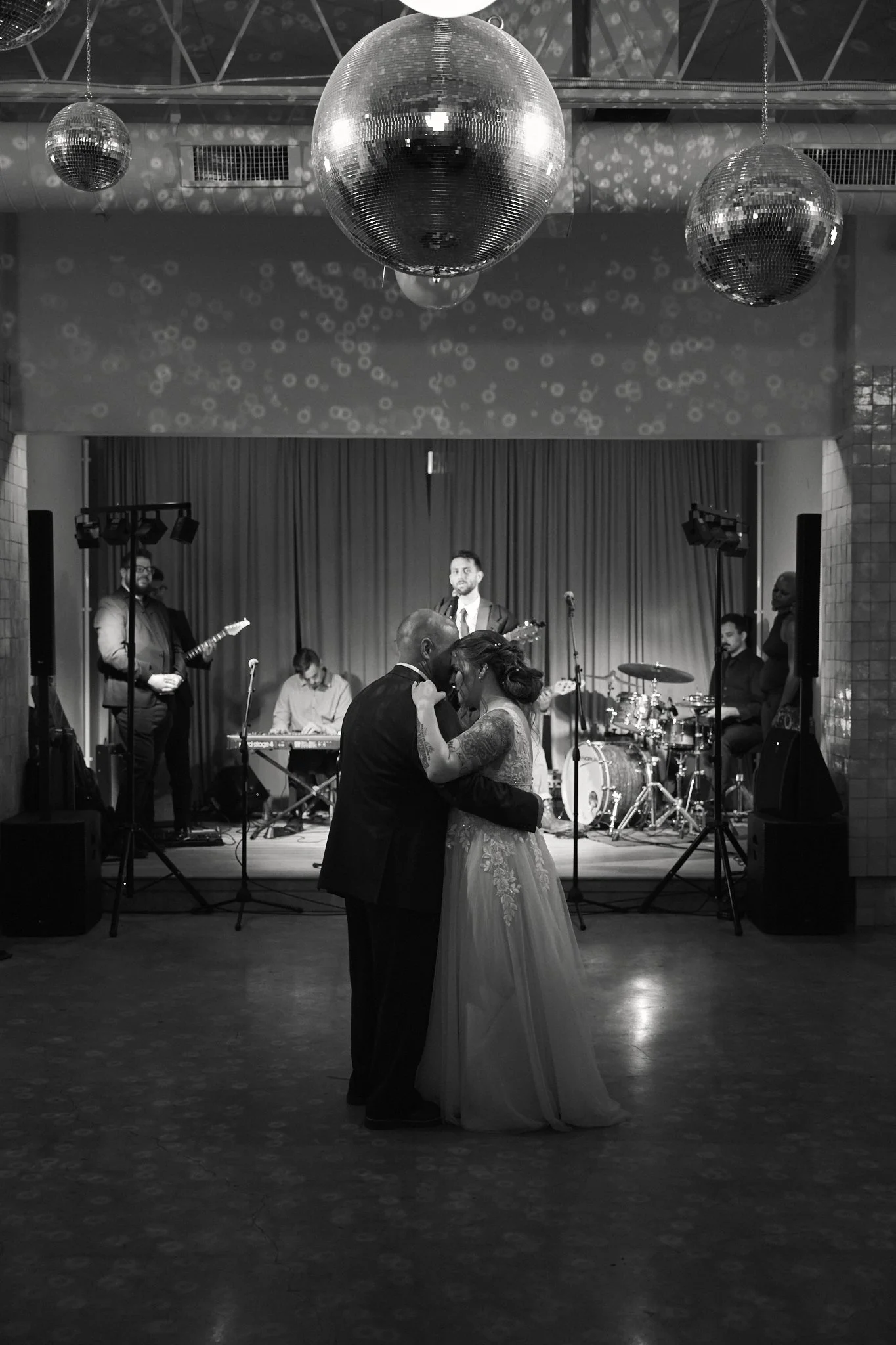 A wedding dance with a newlywed couple dancing closely in the foreground, a band performing live on stage in the background, and disco balls hanging from the ceiling, creating reflections and light patterns on the wall.