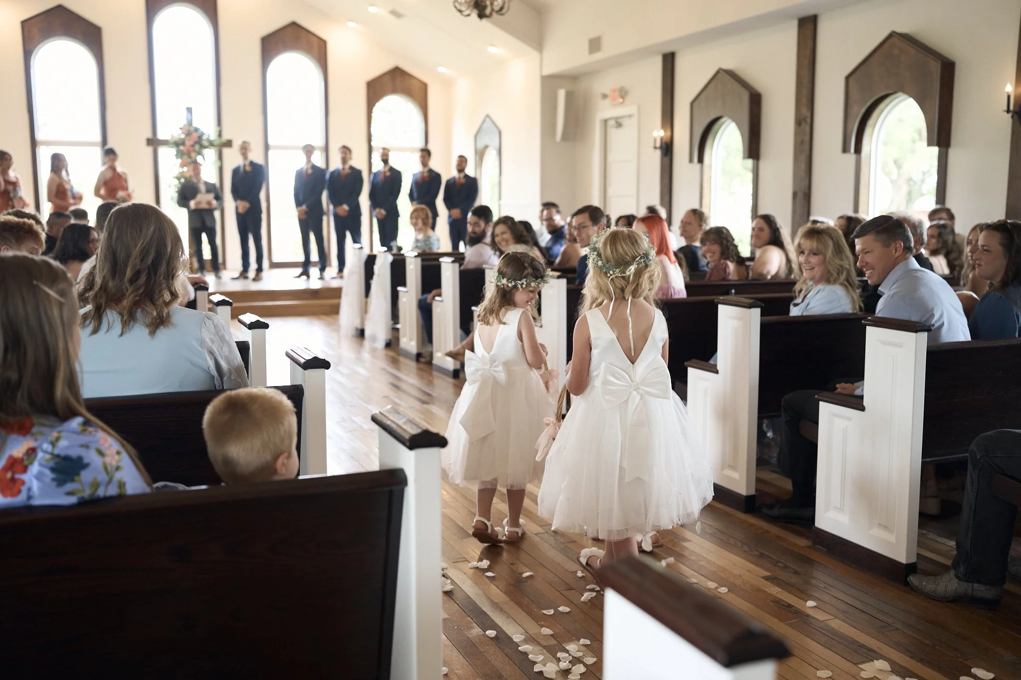 Children dressed in white flower girl dresses holding hands walking down the aisle during a wedding ceremony in a church with guests sitting in pews on either side.