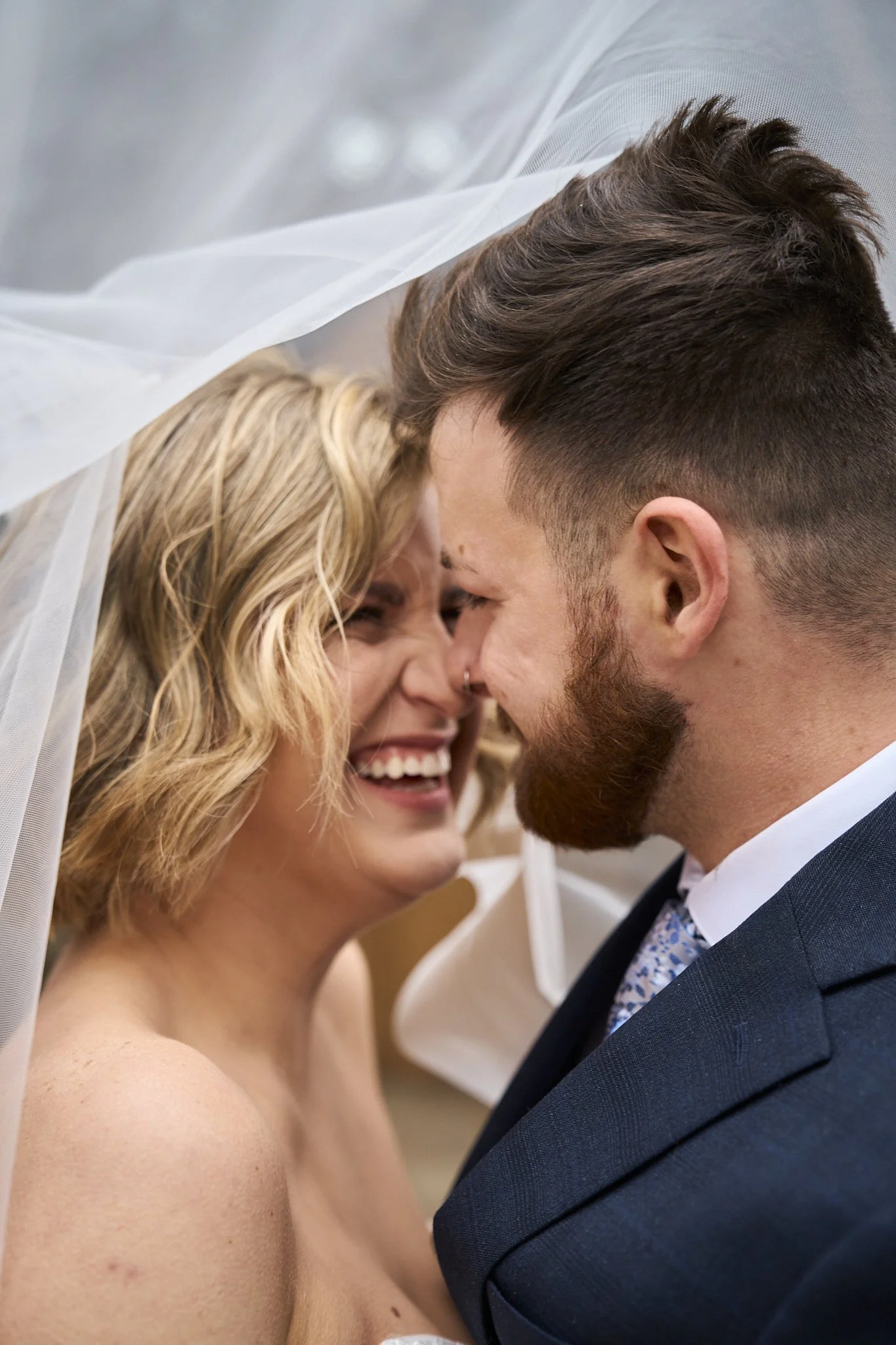 Close-up of a couple with foreheads touching, smiling joyfully during their wedding ceremony. The bride is topless, with blonde curly hair, and the groom is wearing a dark suit and tie.