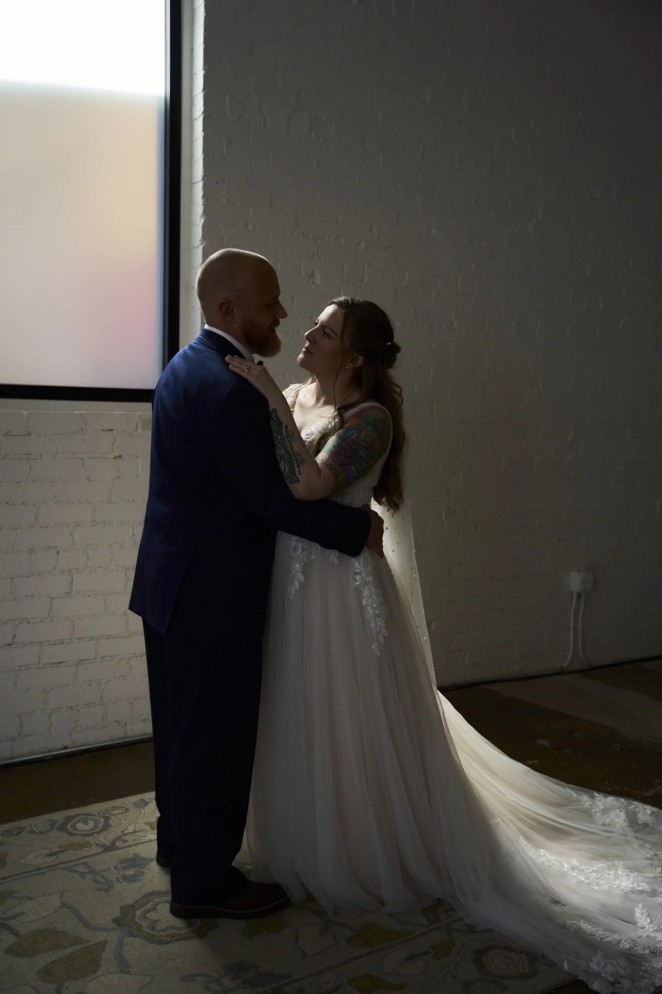 A bride and groom dancing during their wedding, with the bride wearing a white wedding gown and the groom in a dark suit, in a room with a white brick wall and a large window.