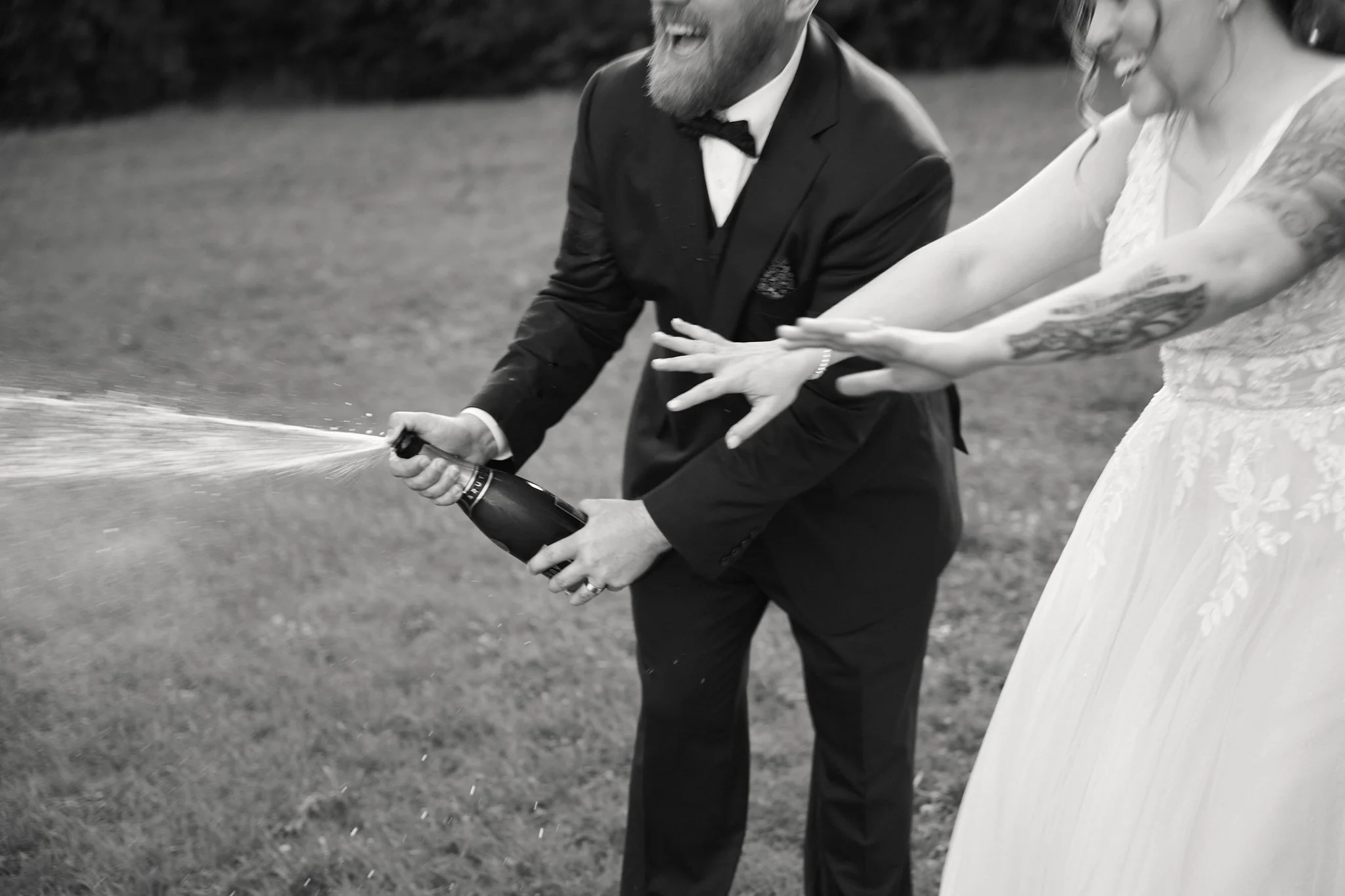 A groom wearing a tuxedo and a bride in a wedding dress are celebrating as they pop a champagne bottle outdoors; the groom is holding the bottle while the bride is reaching towards it, both smiling.