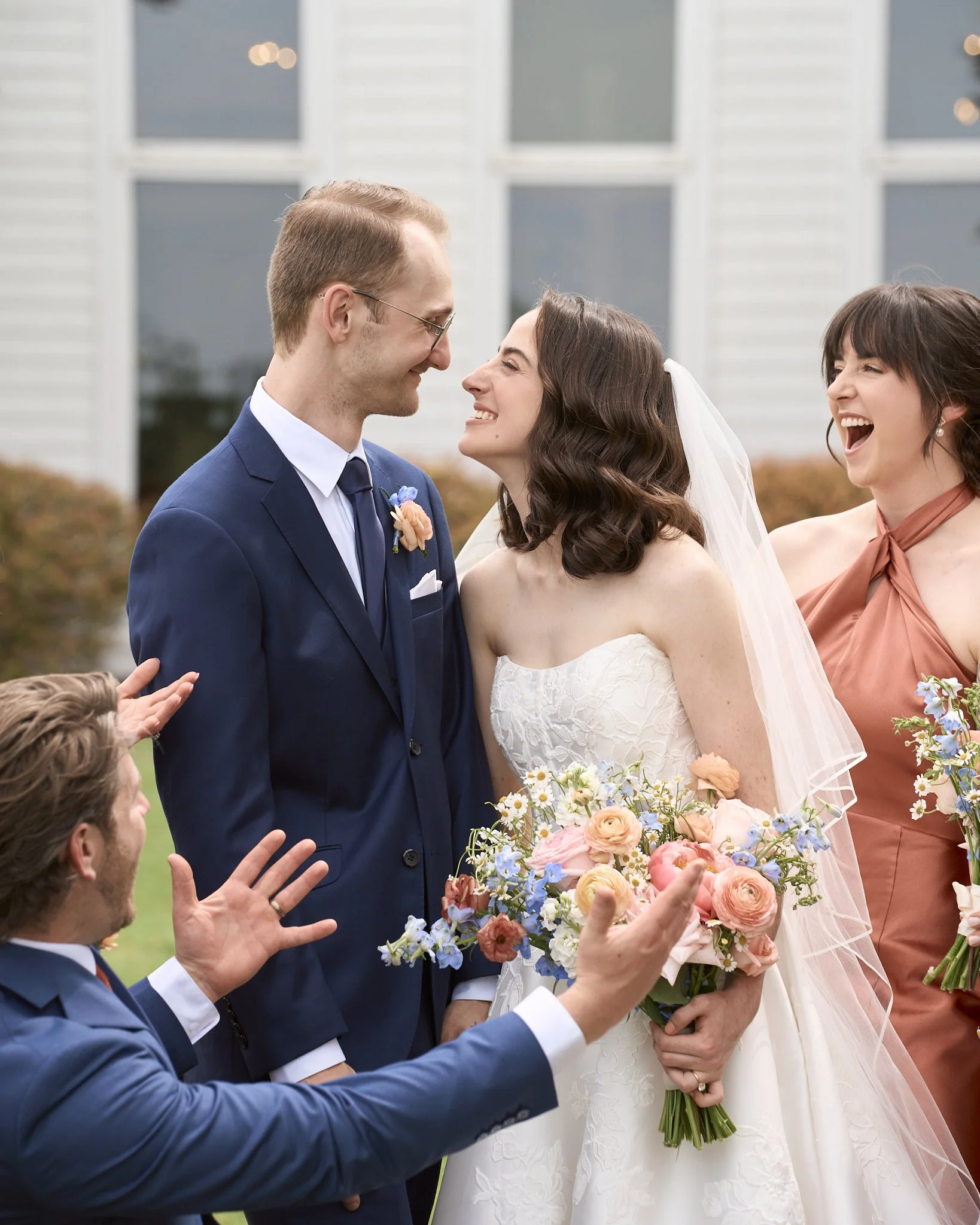 A wedding scene with a bride and groom smiling at each other, holding a bouquet of flowers, while officiants or guests in suits are gesturing and celebrating outdoors.