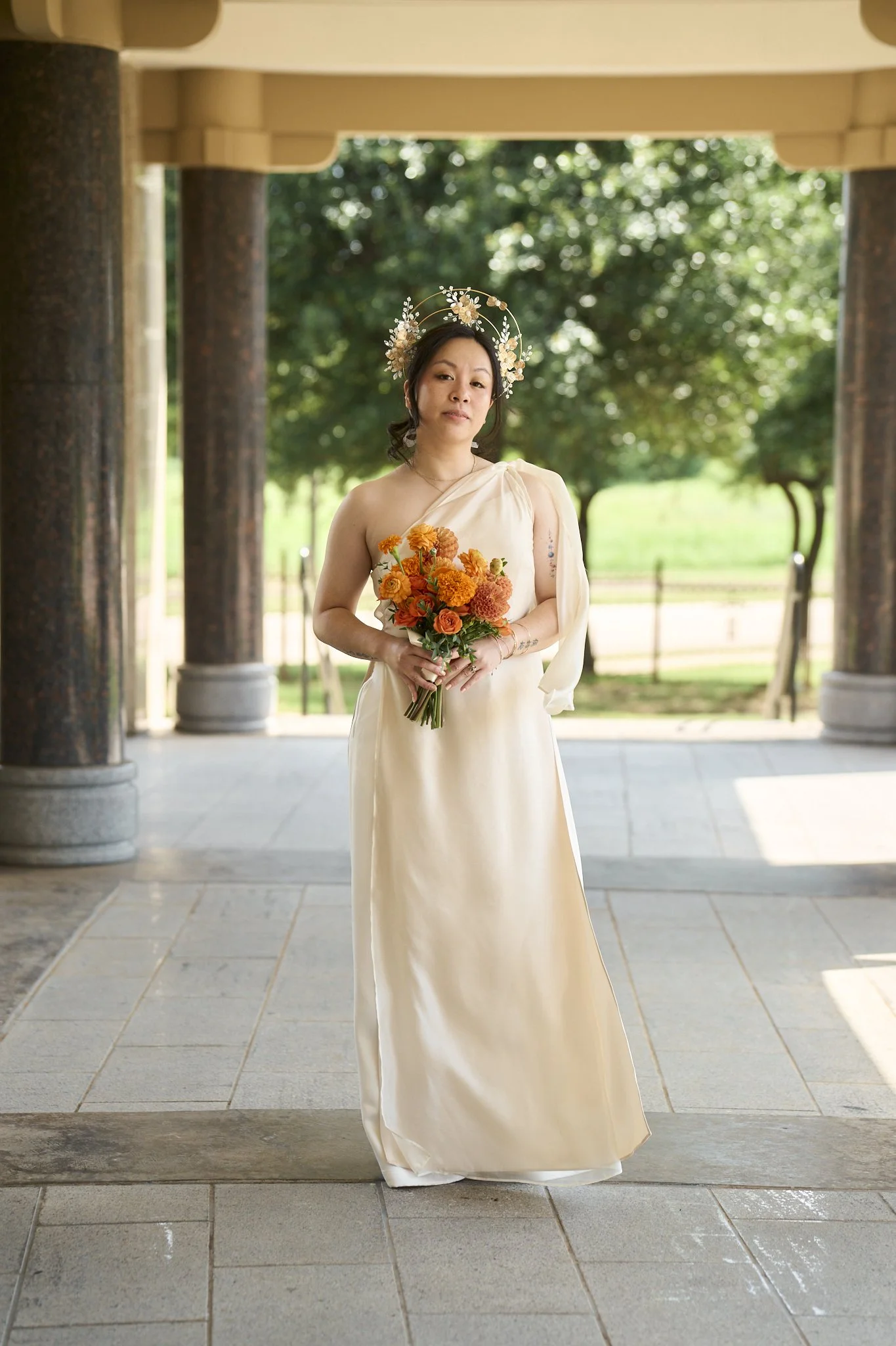 A woman wearing a cream-colored dress with one shoulder strap, holding a bouquet of orange and peach flowers, standing beneath a pergola with tall columns. She has a floral headpiece and stands outdoors with trees and a green landscape in the backgro