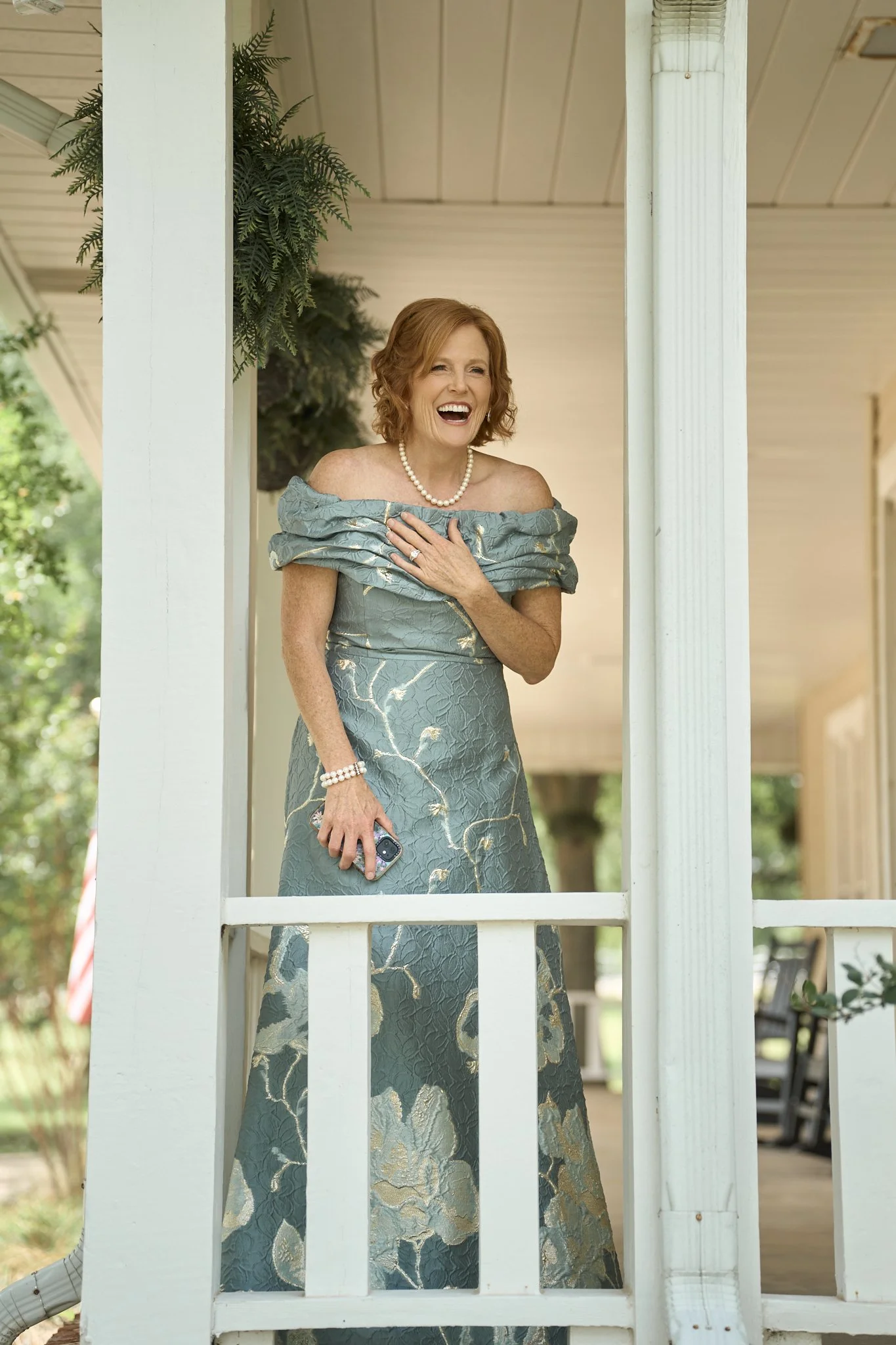 A red-headed woman in an off-the-shoulder blue gown with gold patterns is standing behind a white wooden porch railing, laughing and holding a smartphone in her right hand. She is wearing a pearl necklace and bracelet, and has her left hand over her 
