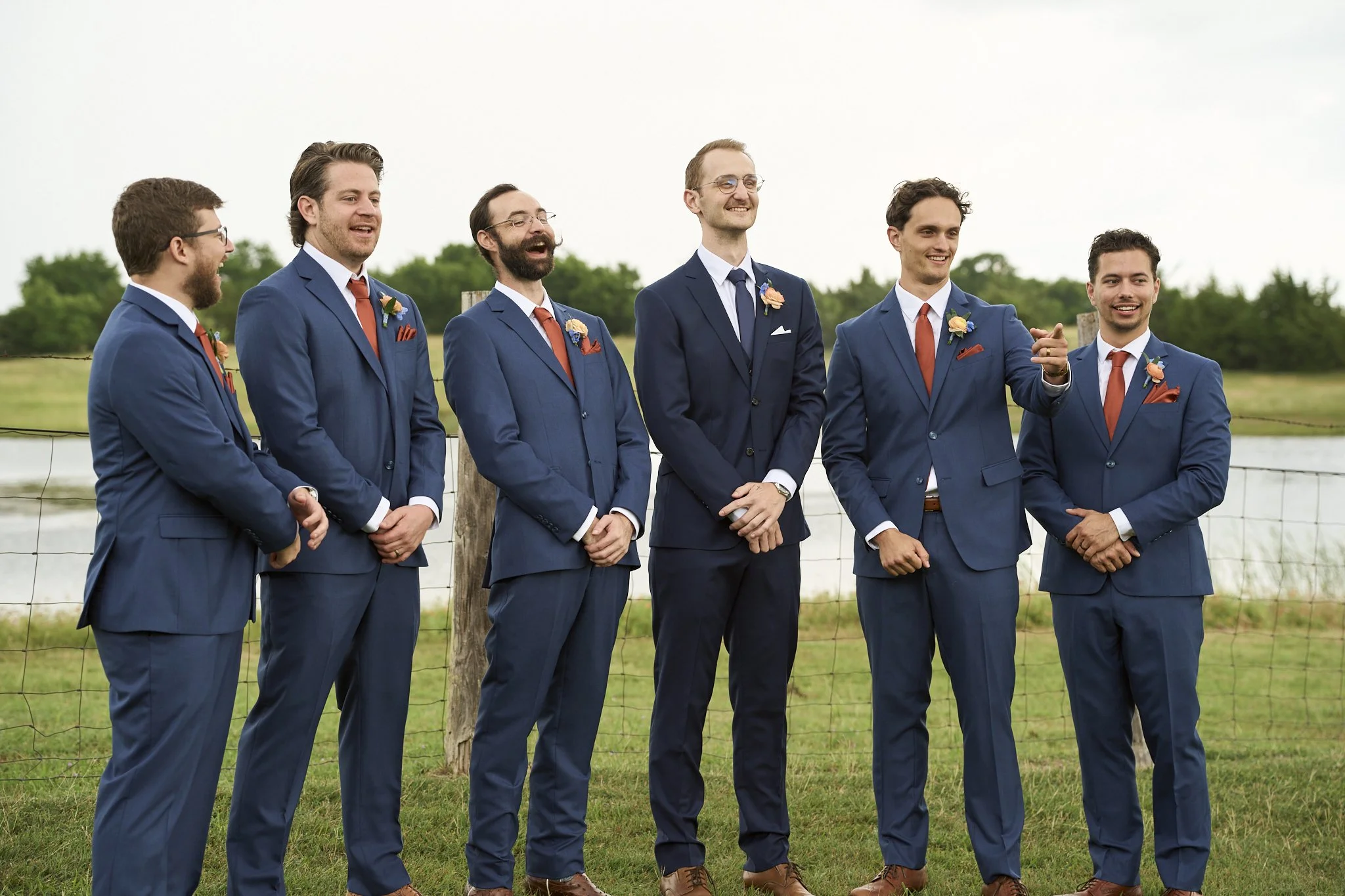 Six men in matching blue suits and orange ties stand outdoors near a wire fence, smiling and laughing. They are dressed formally, with small flowers on their lapels. The background features a cloudy sky and green trees.