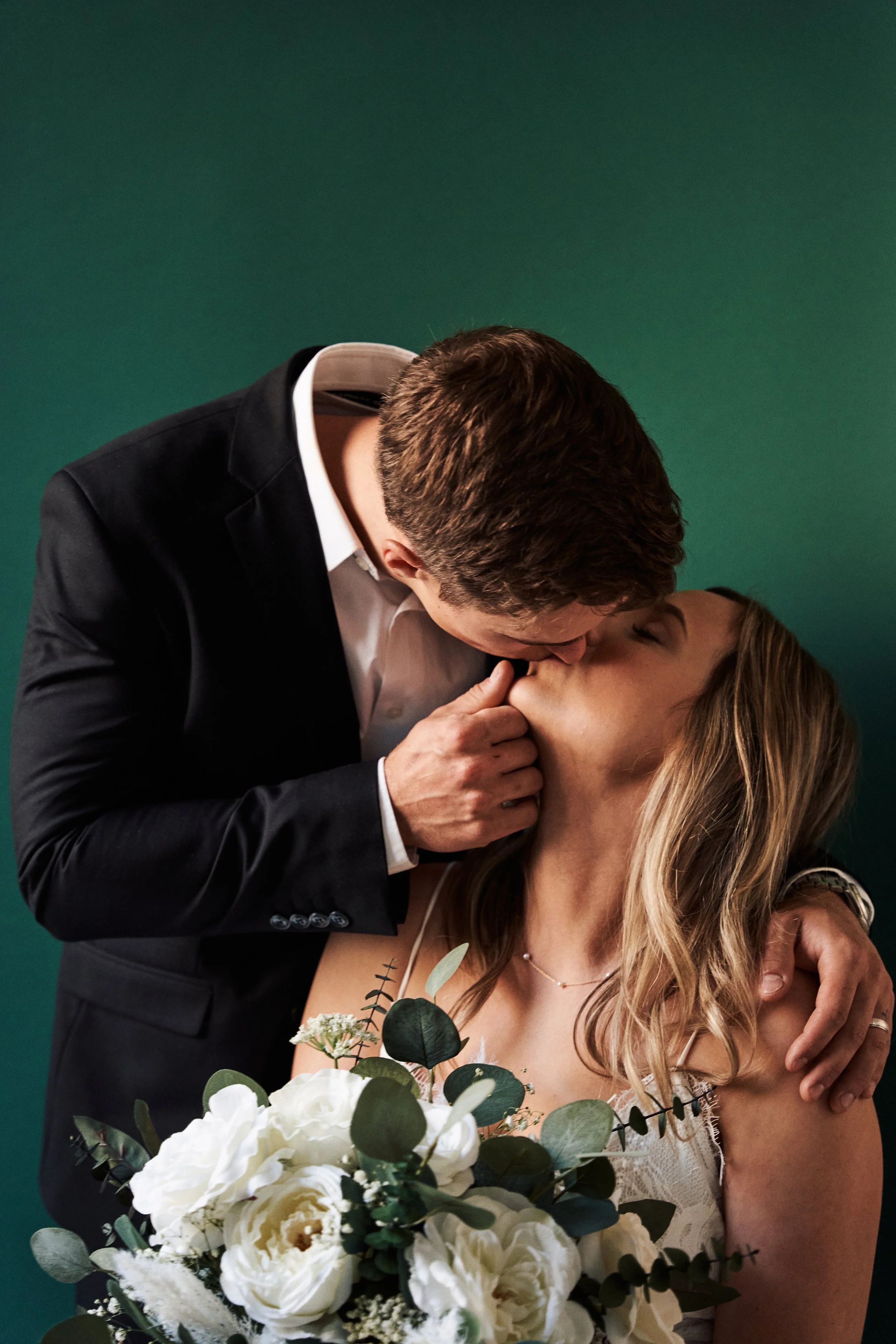 A couple is kissing, the man in a black suit and the woman in a white dress holding a bouquet of white flowers with green leaves. The background is a solid dark green.