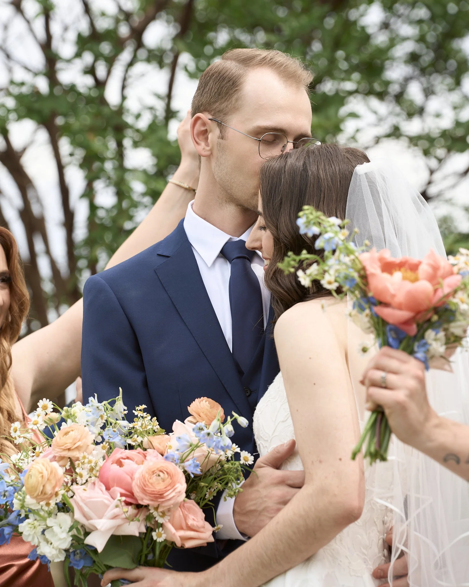 A man and woman, likely at a wedding, are kissing with eyes closed outdoors. The man is wearing glasses, a suit, and a tie, and is holding a bouquet of pink, peach, and blue flowers. The woman has long dark hair, a veil, and is holding a bouquet of s