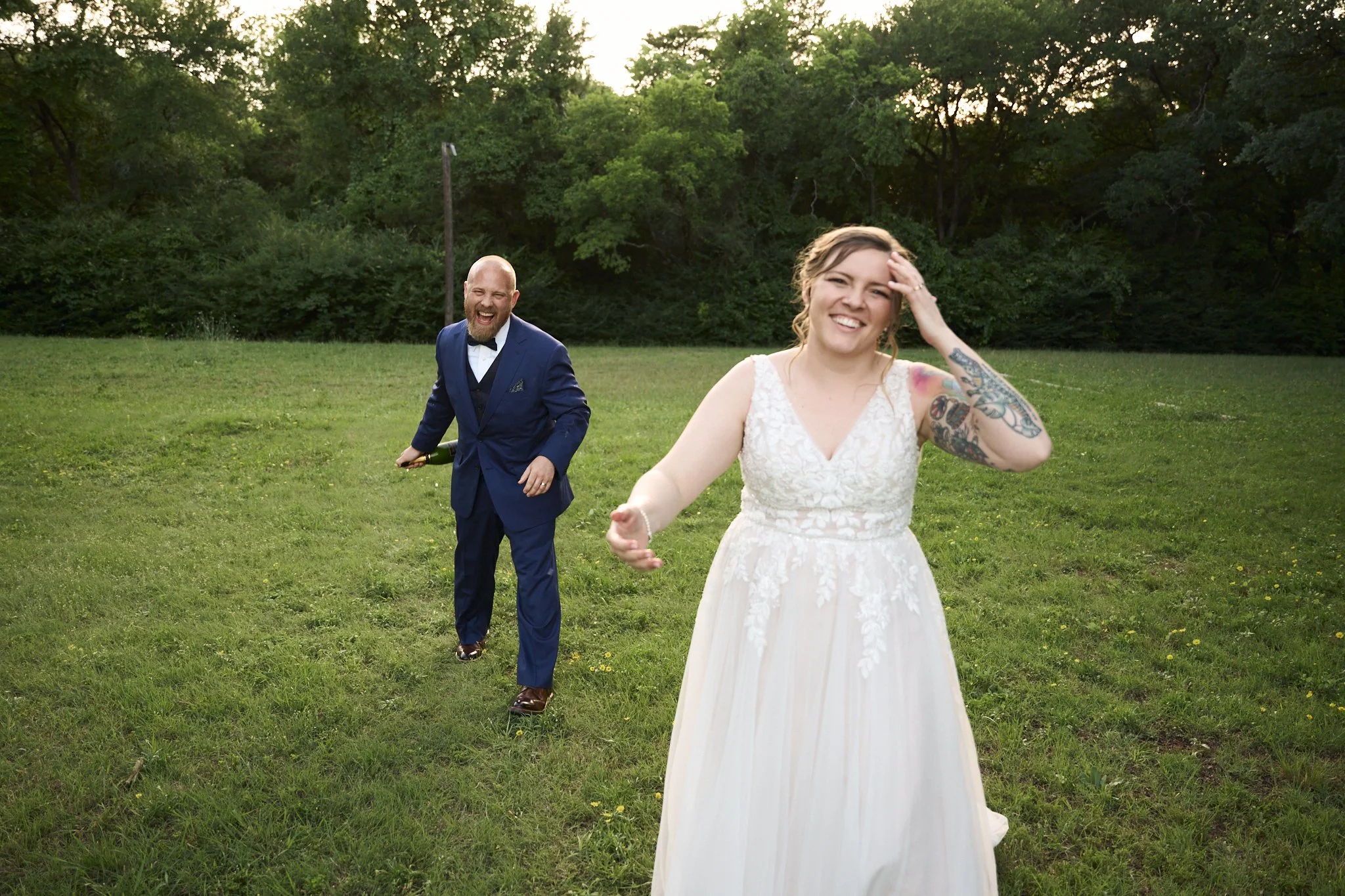 A smiling bride in a white lace wedding dress with tattoos on her arm, and a happy groom in a blue suit with a bow tie, walking on a grassy field with trees in the background.
