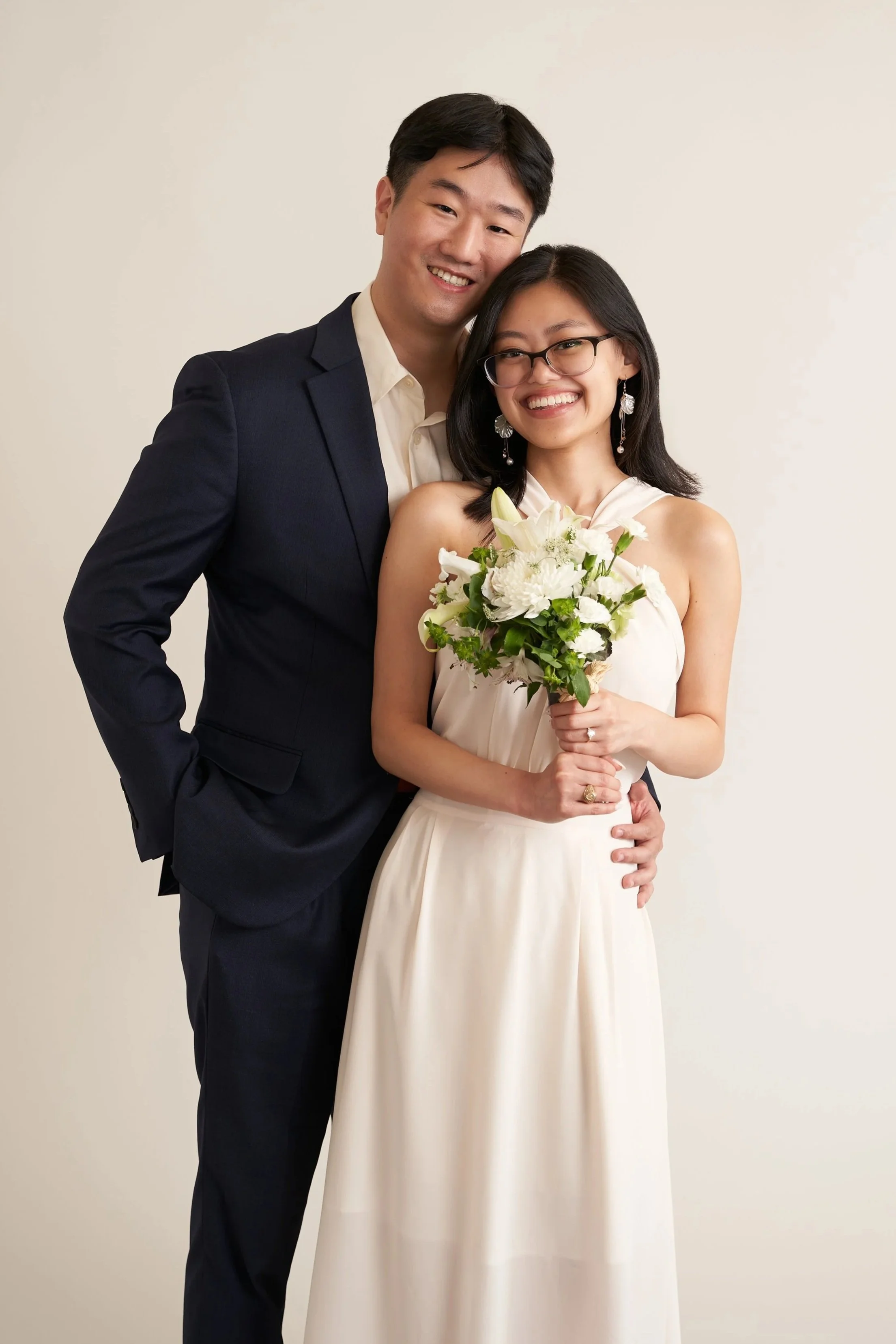 A smiling man in a dark suit standing close to a woman in a cream-colored dress, holding a bouquet of white flowers, both posing against a plain light background.