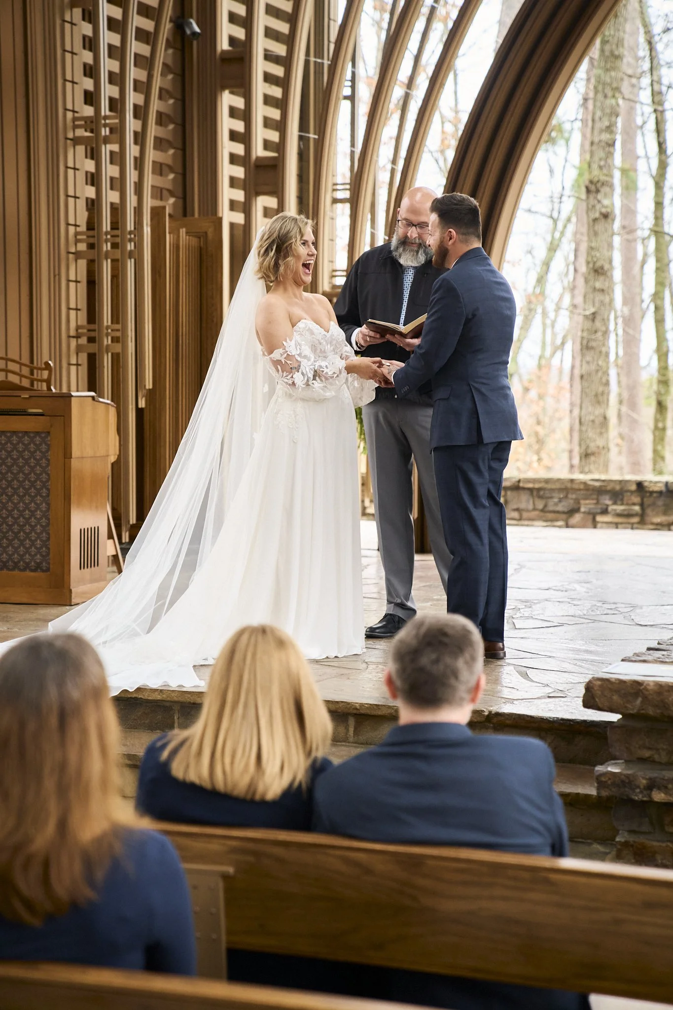 A wedding ceremony taking place in a wooden church with a happy bride and groom holding hands, standing in front of a minister who is reading from a book. The bride is in a white dress with lace details, and the groom is in a dark suit. Guests are se