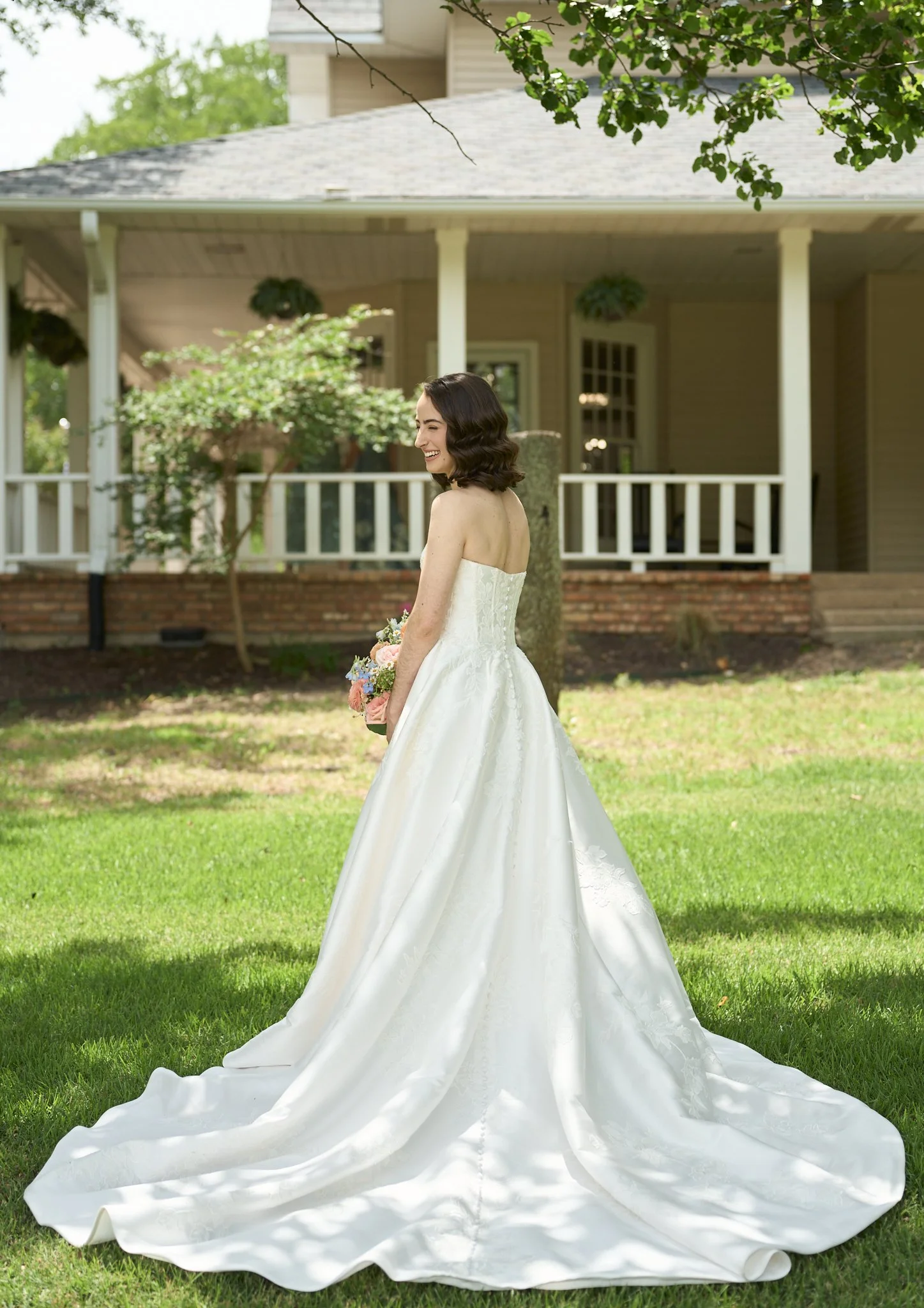 A smiling woman in a white wedding gown holding a floral bouquet stands on a grassy lawn in front of a house with a porch, trees, and hanging plants.