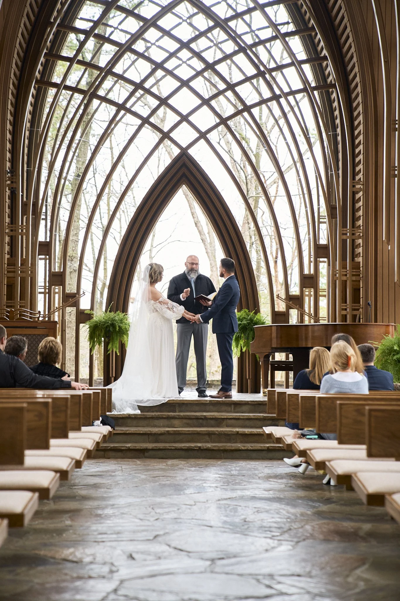 A wedding ceremony taking place inside a church with wooden gothic-style architecture. A couple is holding hands in front of an officiant, with the bride wearing a white wedding dress and veil, and the groom in a blue suit. Guests are seated on woode