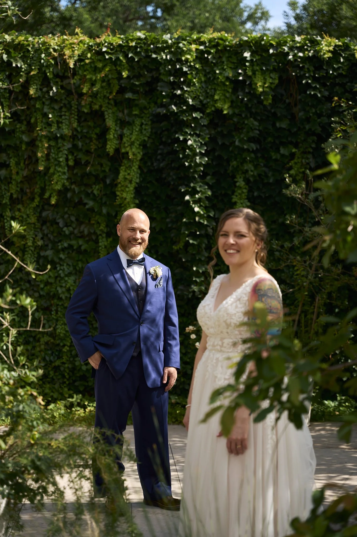 A couple, dressed in wedding attire, standing outdoors in front of a green ivy-covered wall. The man is wearing a blue suit with a bow tie, and the woman is wearing a white lace dress with floral tattoos on her arms.