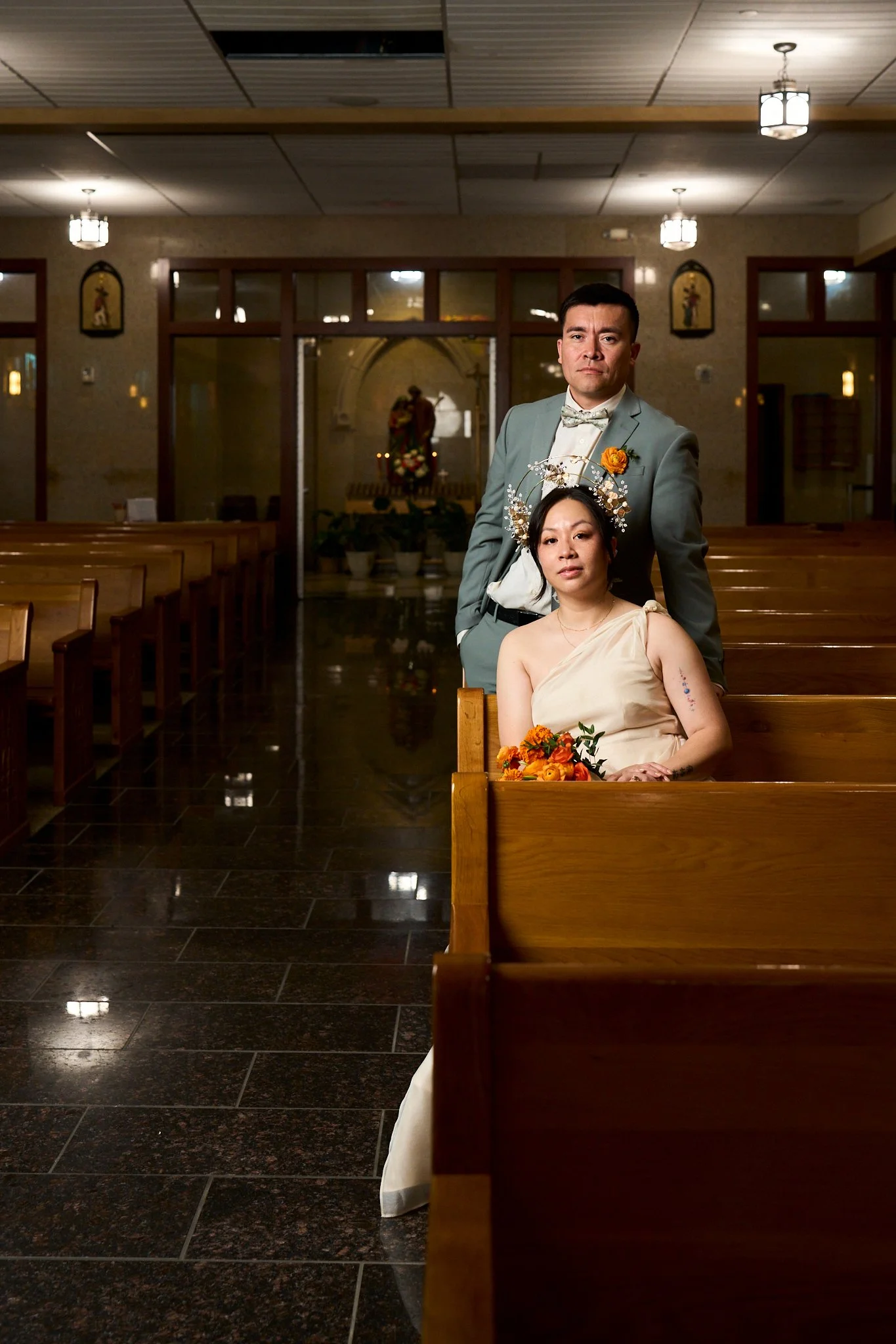 A man and a woman inside a church. The woman is sitting on a pew, wearing a cream-colored dress, a floral headpiece, and holding a bouquet. The man is standing behind her, wearing a light gray suit with a bow tie.