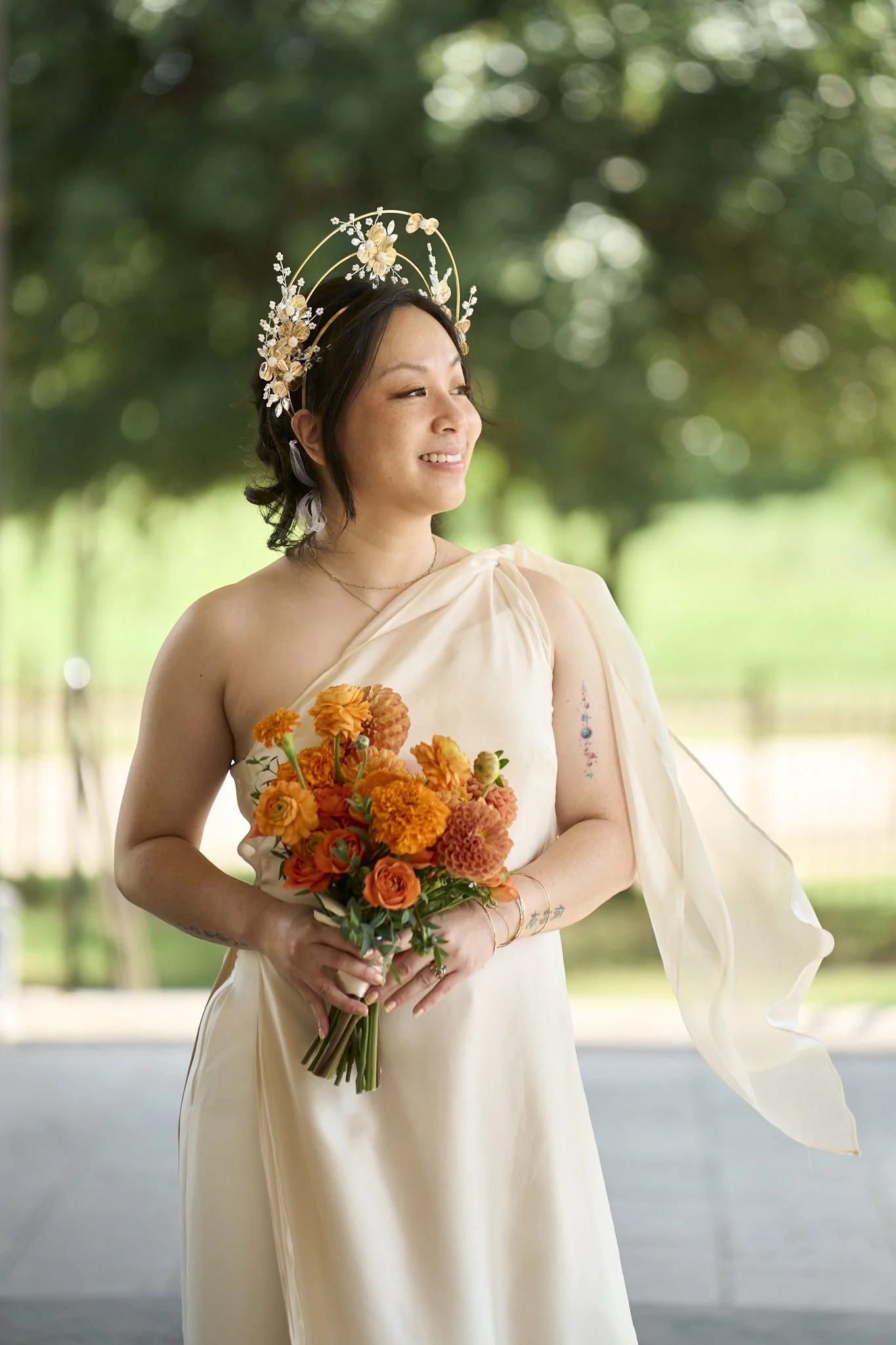 A woman wearing a cream-colored dress with one shoulder and a sheer, flowing sleeve, holding a bouquet of orange and yellow flowers. She has a floral headpiece and tattoos on her arm, standing outdoors with blurred greenery in the background, smiling