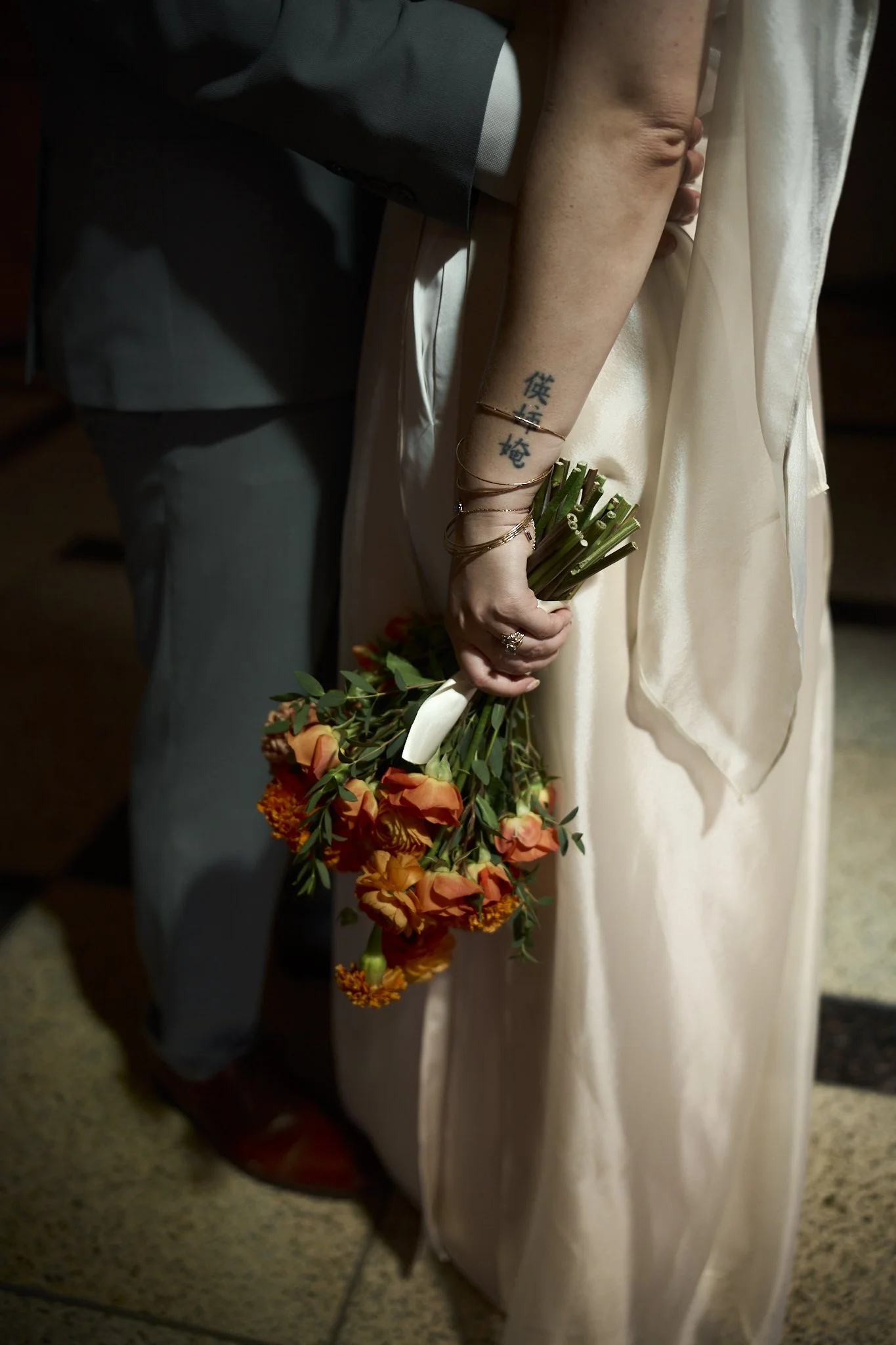 Close-up of a woman's hand holding a bouquet of orange and yellow flowers, with a tattoo of Asian characters on her forearm, and wearing bracelets. She is dressed in a light-colored satin or silk gown, standing next to a person dressed in a dark suit