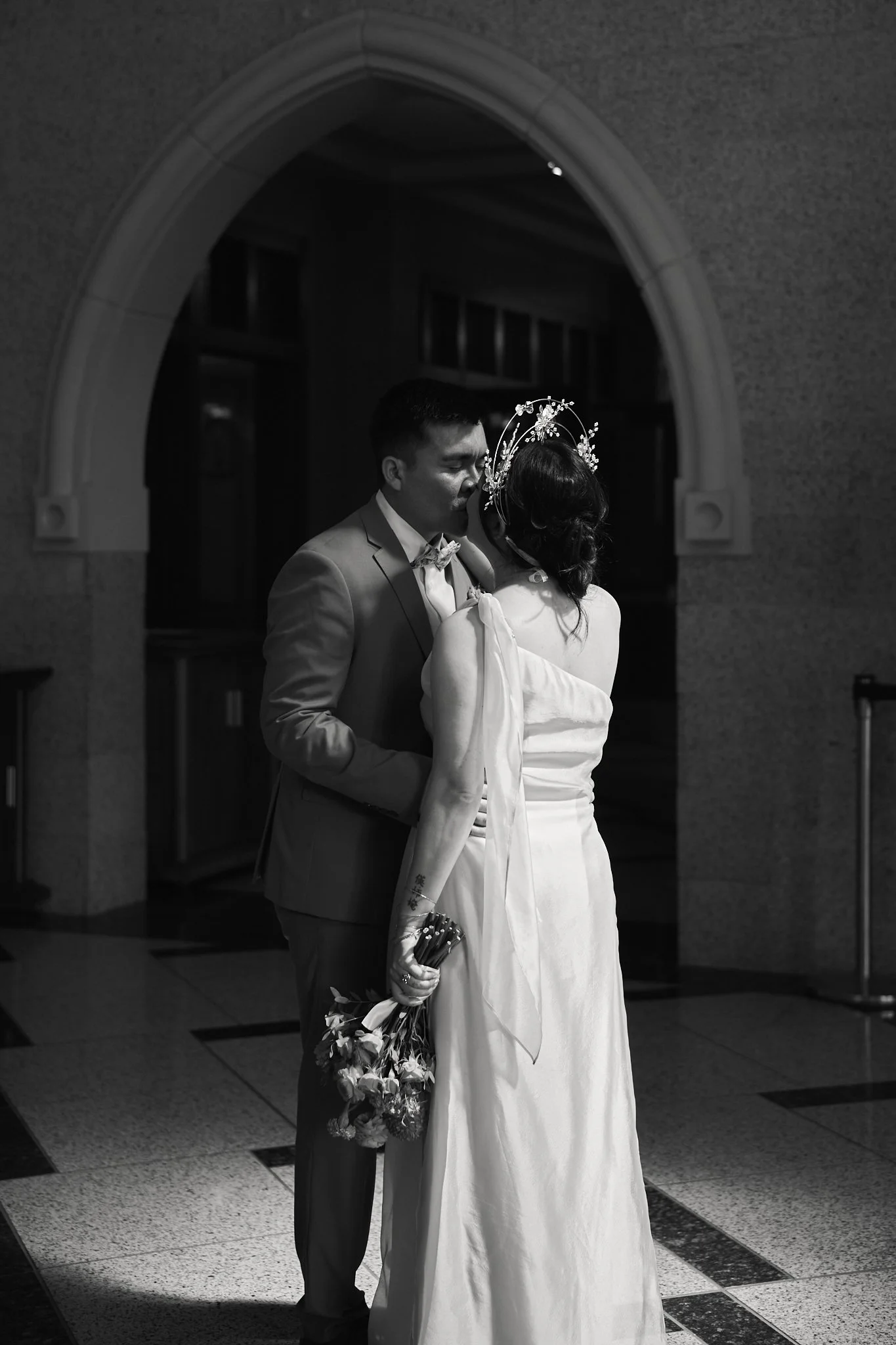 A black and white photo of a couple sharing a kiss in a dimly lit indoor space. The man is dressed in a suit, and the woman is wearing a white dress with a decorative headpiece. The woman is holding a bouquet of flowers.
