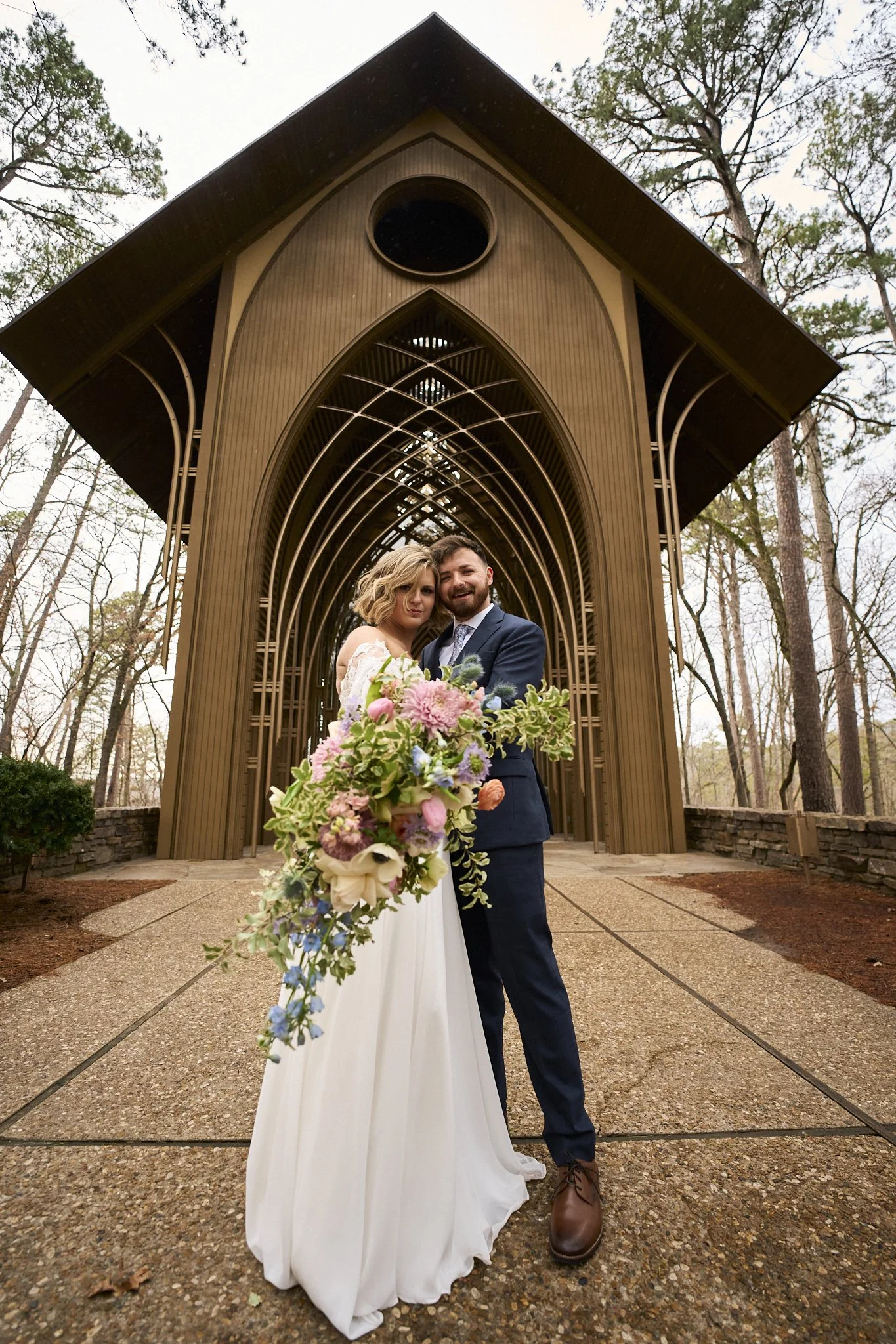A bride and groom standing under a church archway, the bride holding a large bouquet and the groom wearing a dark suit, in an outdoor setting with trees in the background.