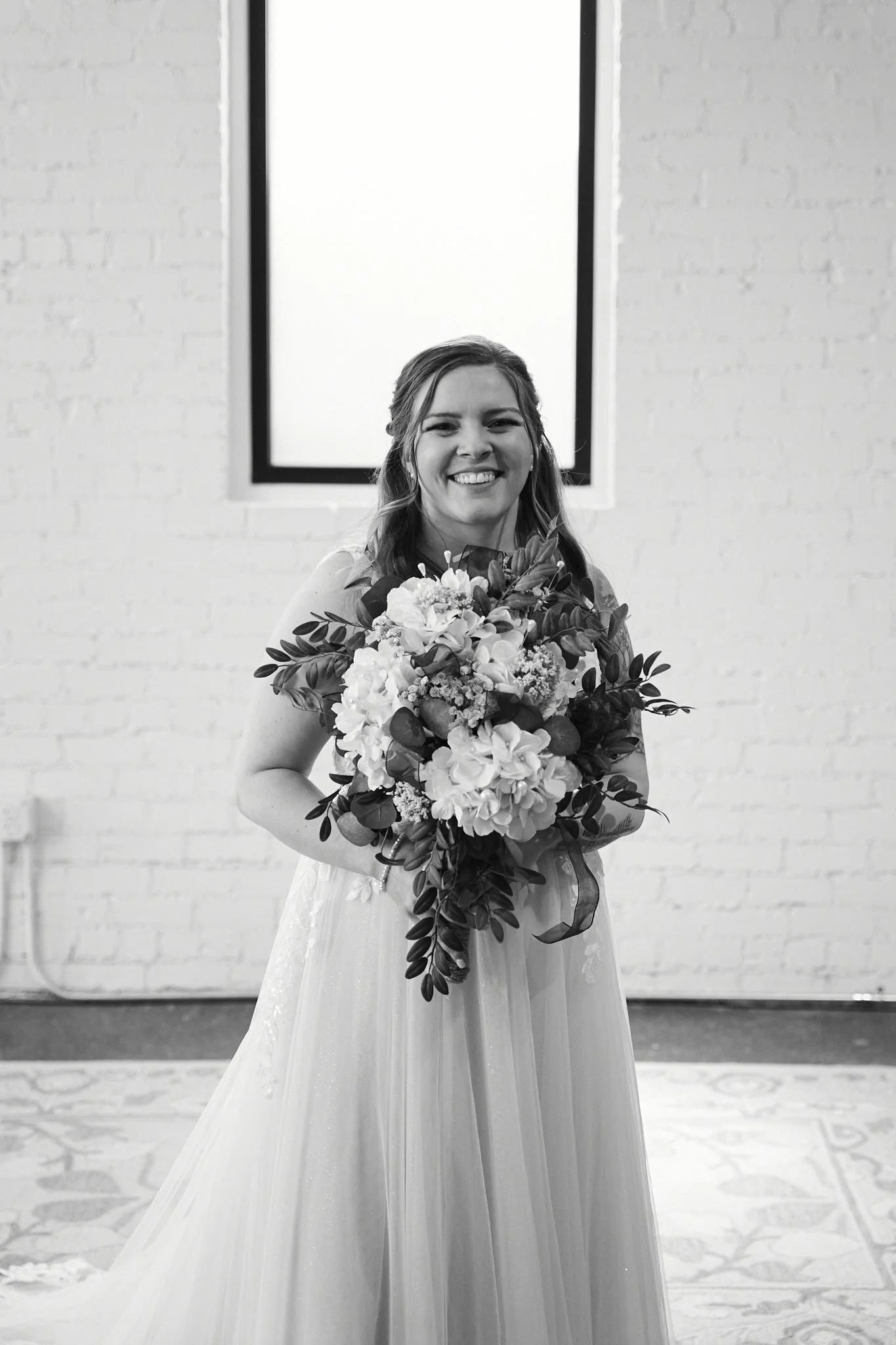 A woman in a wedding dress holding a bouquet of flowers, smiling in front of a white brick wall and a large window.