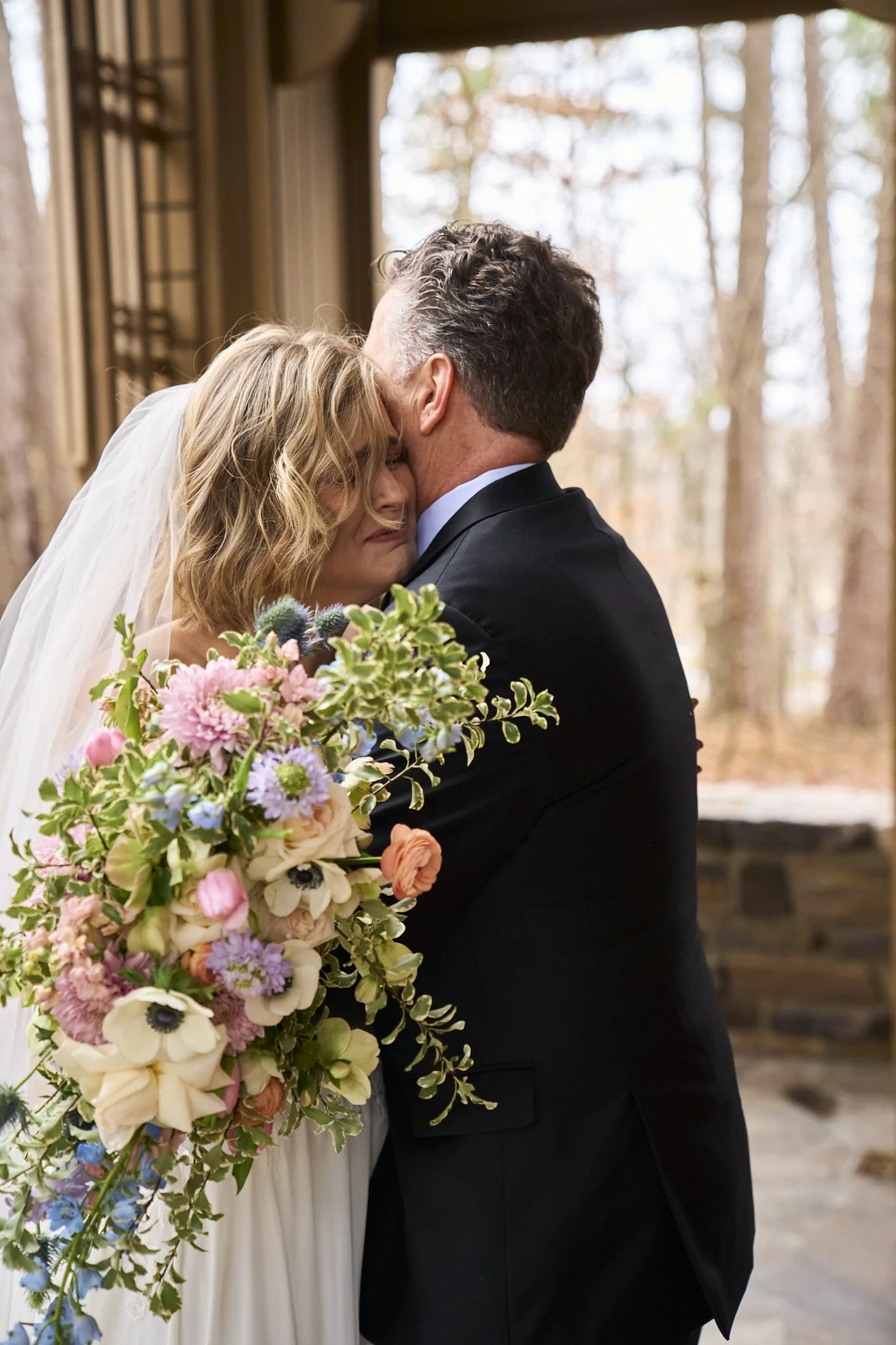 A bride and groom embrace during a wedding ceremony in a wooded outdoor setting, with the bride holding a large colorful bouquet of flowers.