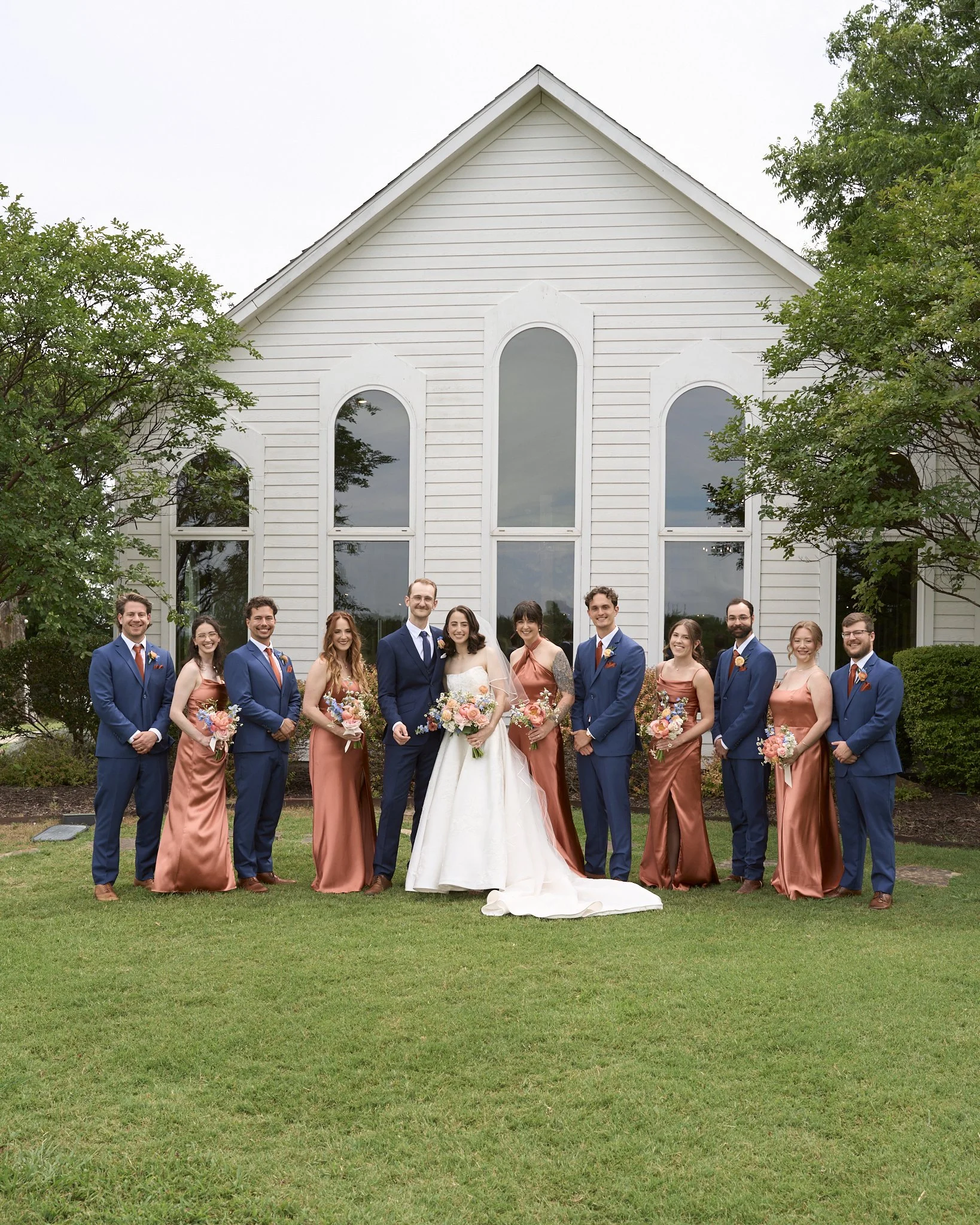 A group of wedding party members, including the bride, groom, bridesmaids, and groomsmen, standing outside in front of a white church building with large arched windows and surrounded by greenery.