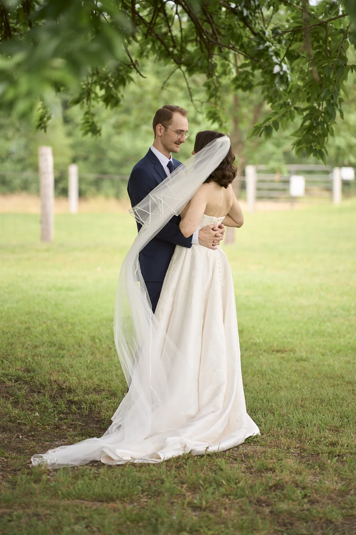 A bride and groom are standing outdoors under a tree, sharing a tender moment on their wedding day. The groom is wearing a navy suit and glasses, and the bride is dressed in a strapless white wedding gown with a long veil.