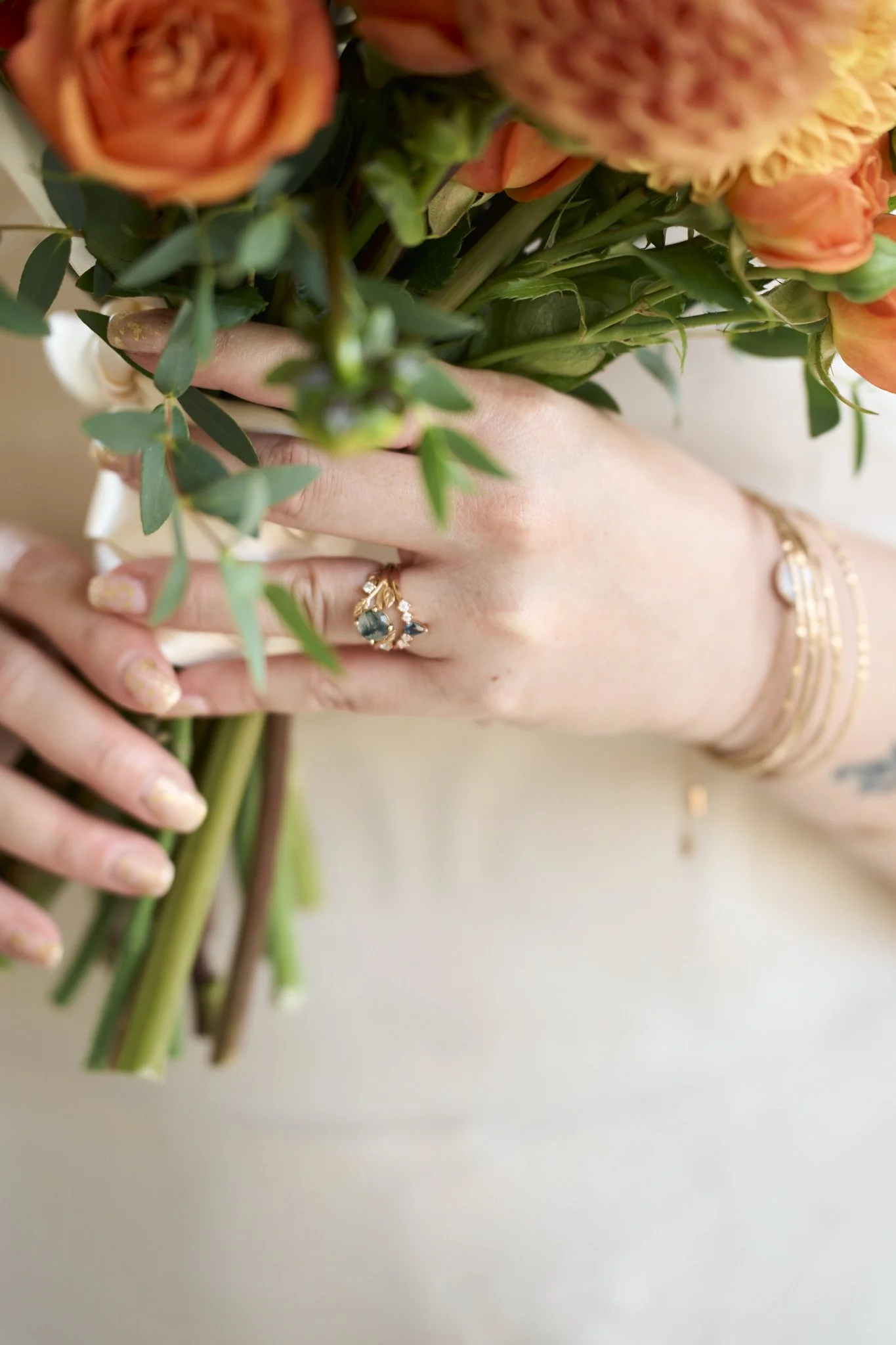 Close-up of a person holding a bouquet of orange and peach-colored flowers with green foliage, wearing rings and gold bracelets.