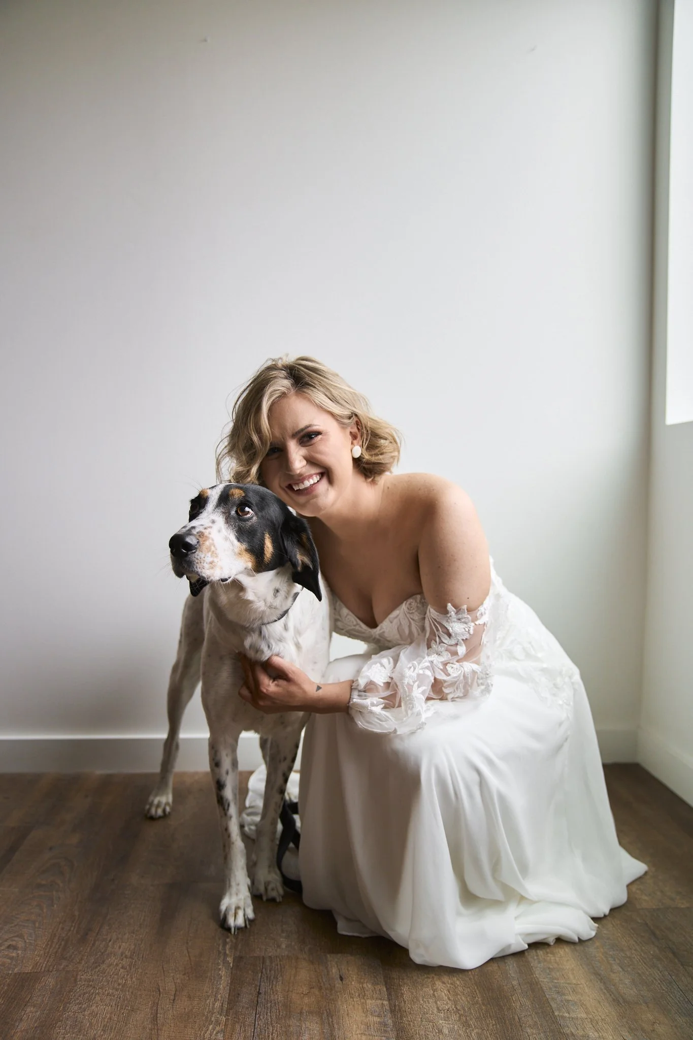 A woman dressed in a white wedding gown kneeling on a hardwood floor, smiling, with her arm around a large black, white, and tan Great Dane dog in a bright room with white walls and a window on the right.