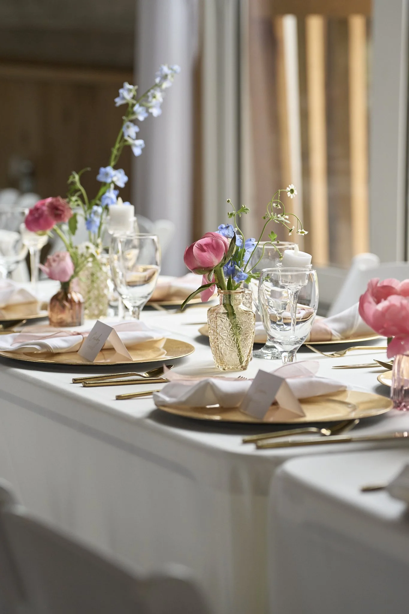 Decorated dining table with pink and blue flowers in glass vases, gold placemats, white napkins, and glassware in a well-lit room.