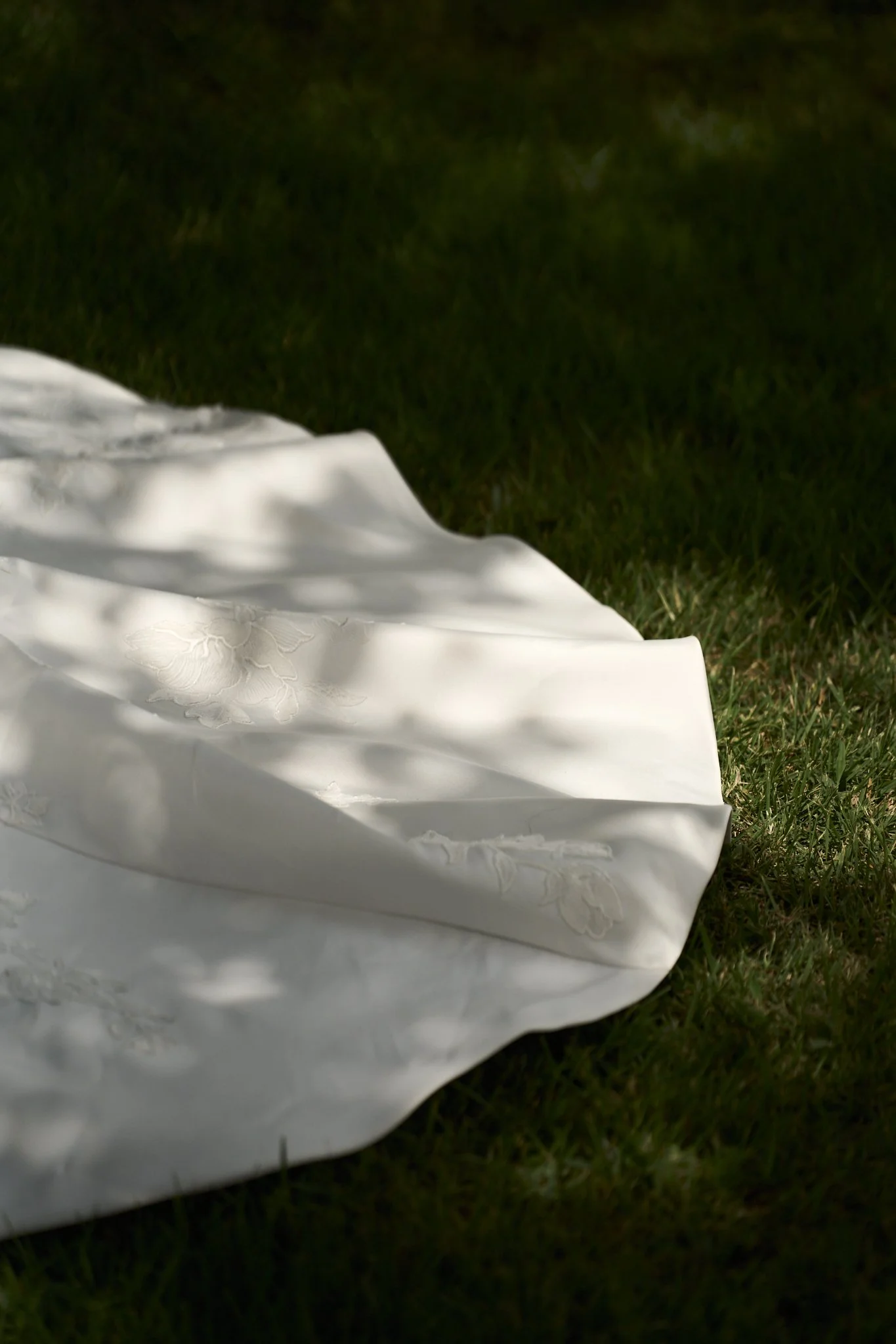 Close-up of a white pillow with embroidered floral design, placed on green grass, shadowed by trees.