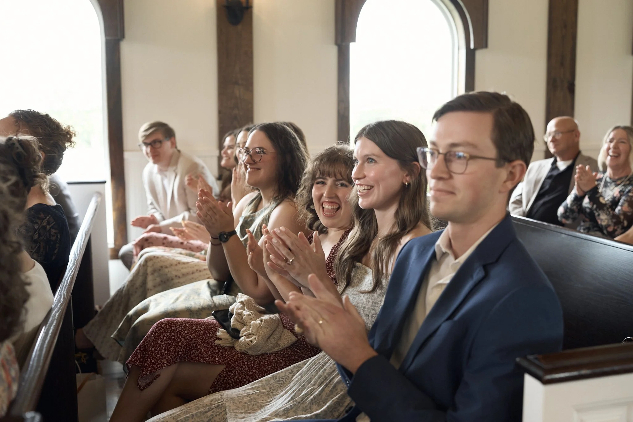 People sitting in pews, clapping, smiling, and laughing during a gathering or event in a church or similar setting.