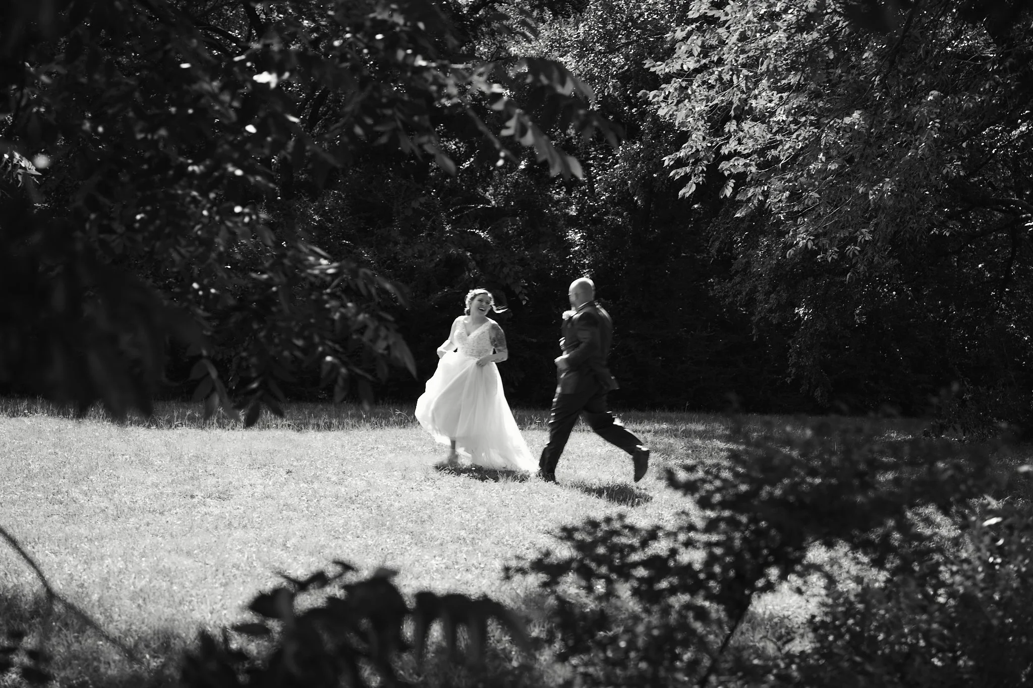 A black and white photo of a bride and groom walking outdoors in a wooded area, with the bride in a wedding dress and the groom in a suit, framed by tree branches.