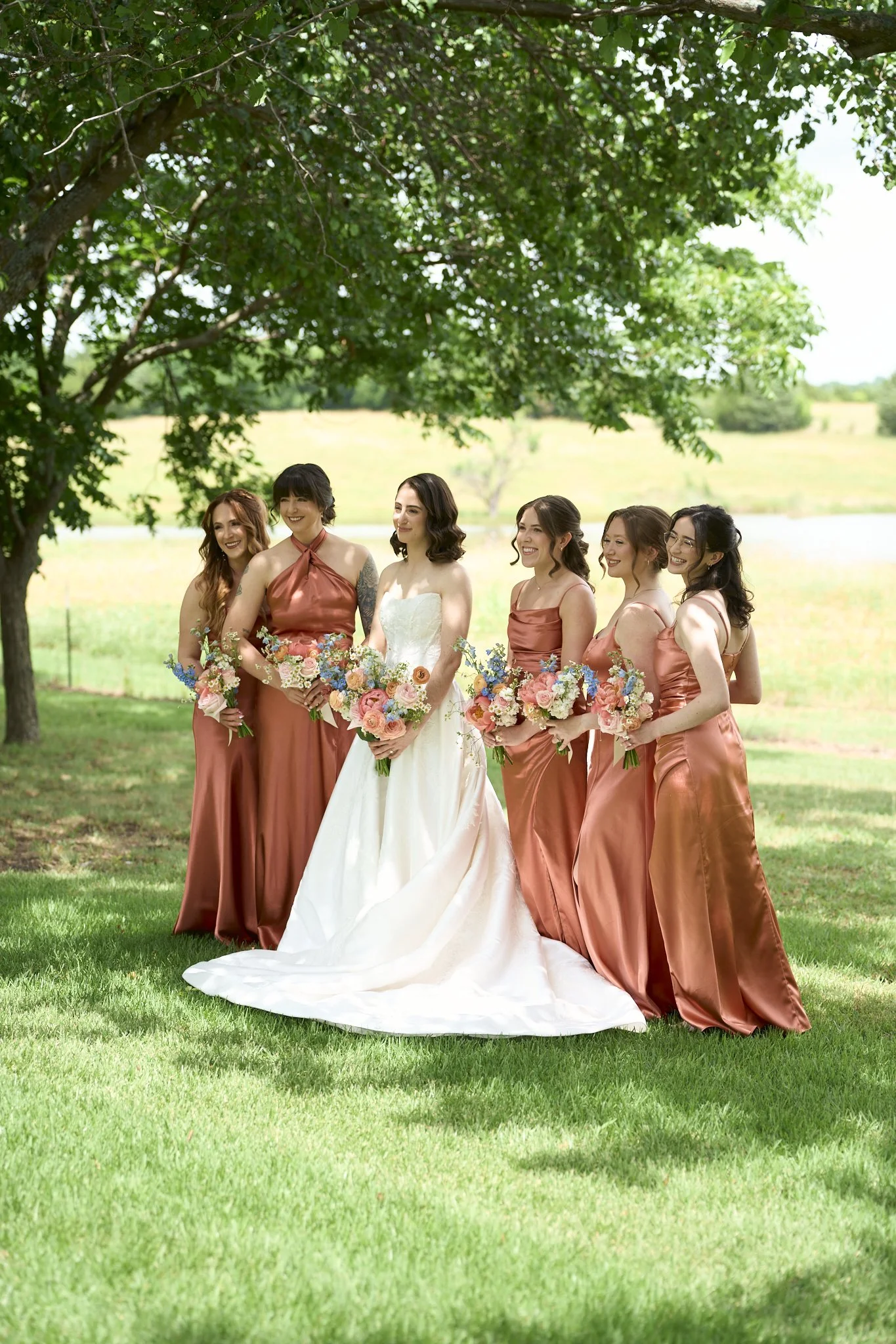 A bride and five bridesmaids standing outdoors under a large leafy tree, holding bouquets of flowers, smiling. The bride is in a white gown, while the bridesmaids are in matching copper-colored dresses.