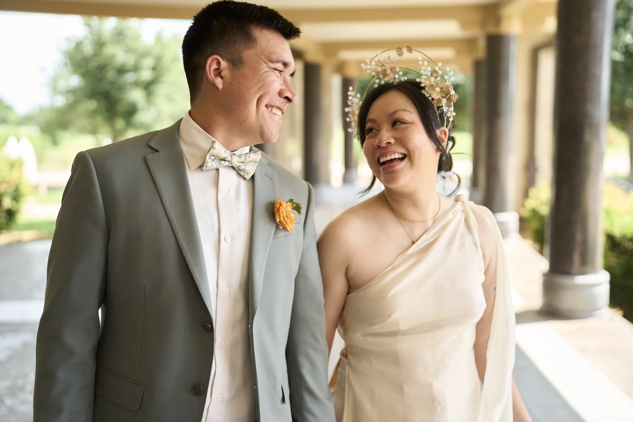 A smiling couple at a wedding, the man in a light gray suit with a floral bow tie and the woman in a cream-colored dress with a floral headpiece, standing beneath a structure with columns and greenery background.