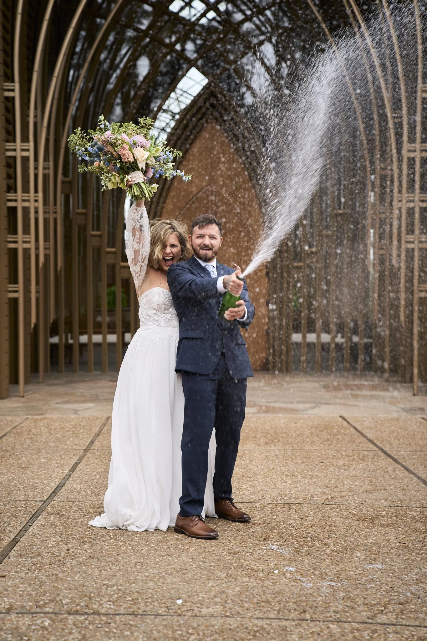 A newlywed couple celebrating outdoors, with the groom spraying champagne and the bride holding a bouquet of flowers