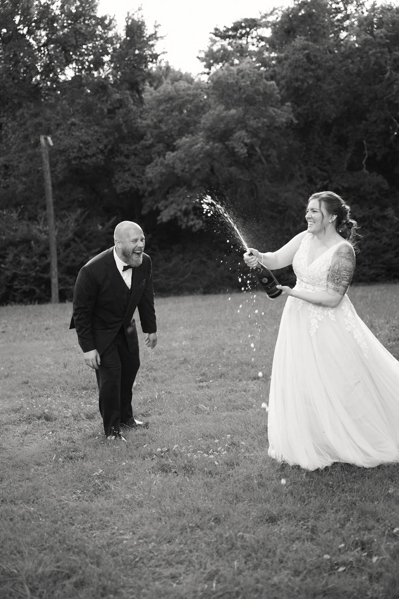A bride and groom celebrate outdoors, with the bride spraying champagne at the groom and both laughing joyfully, in a grassy field with trees in the background.