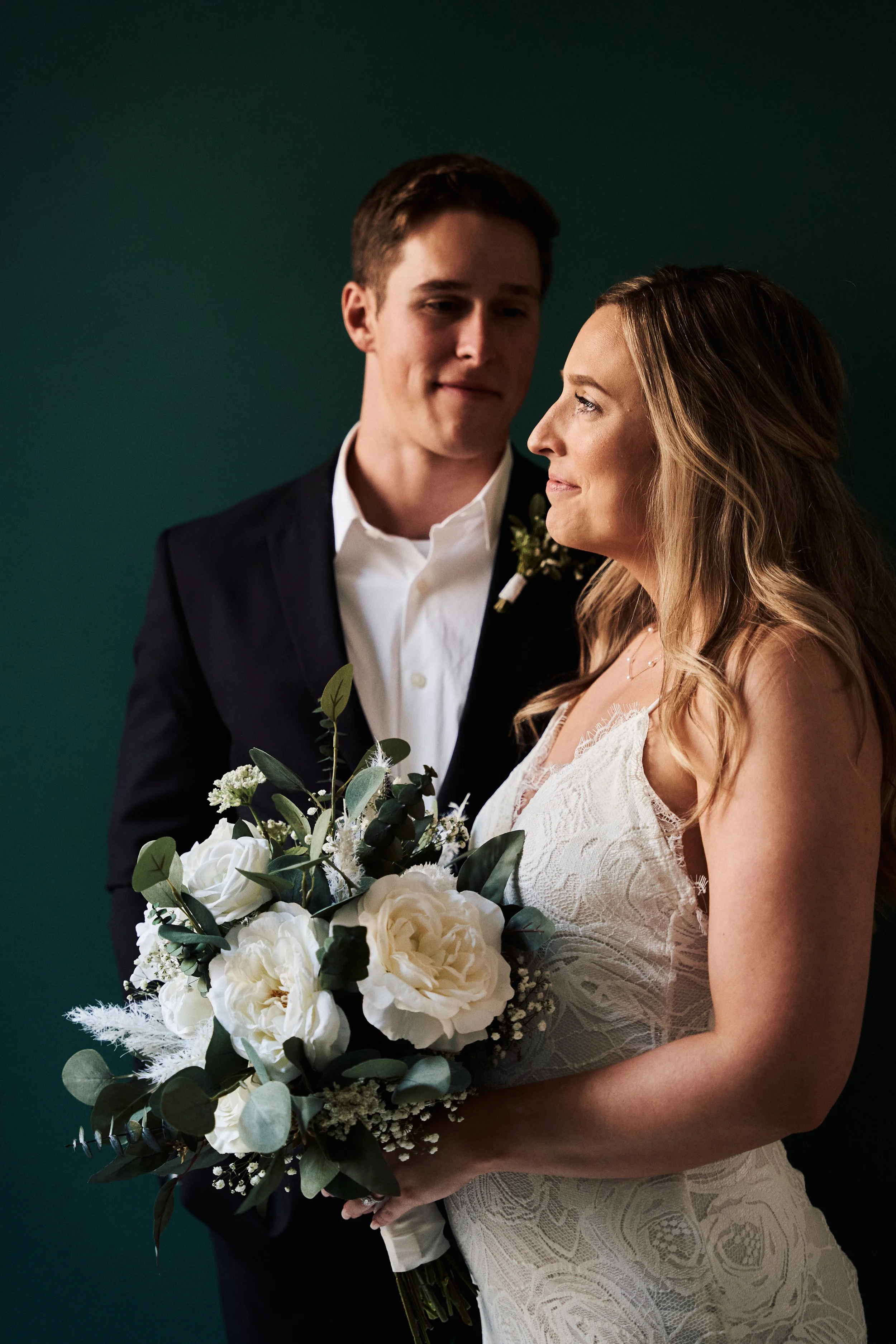 A bride and groom standing close together, with the bride holding a bouquet of white flowers and greenery. The bride wears a lace wedding dress, and the groom is dressed in a black suit with a white shirt. They are looking at each other affectionatel