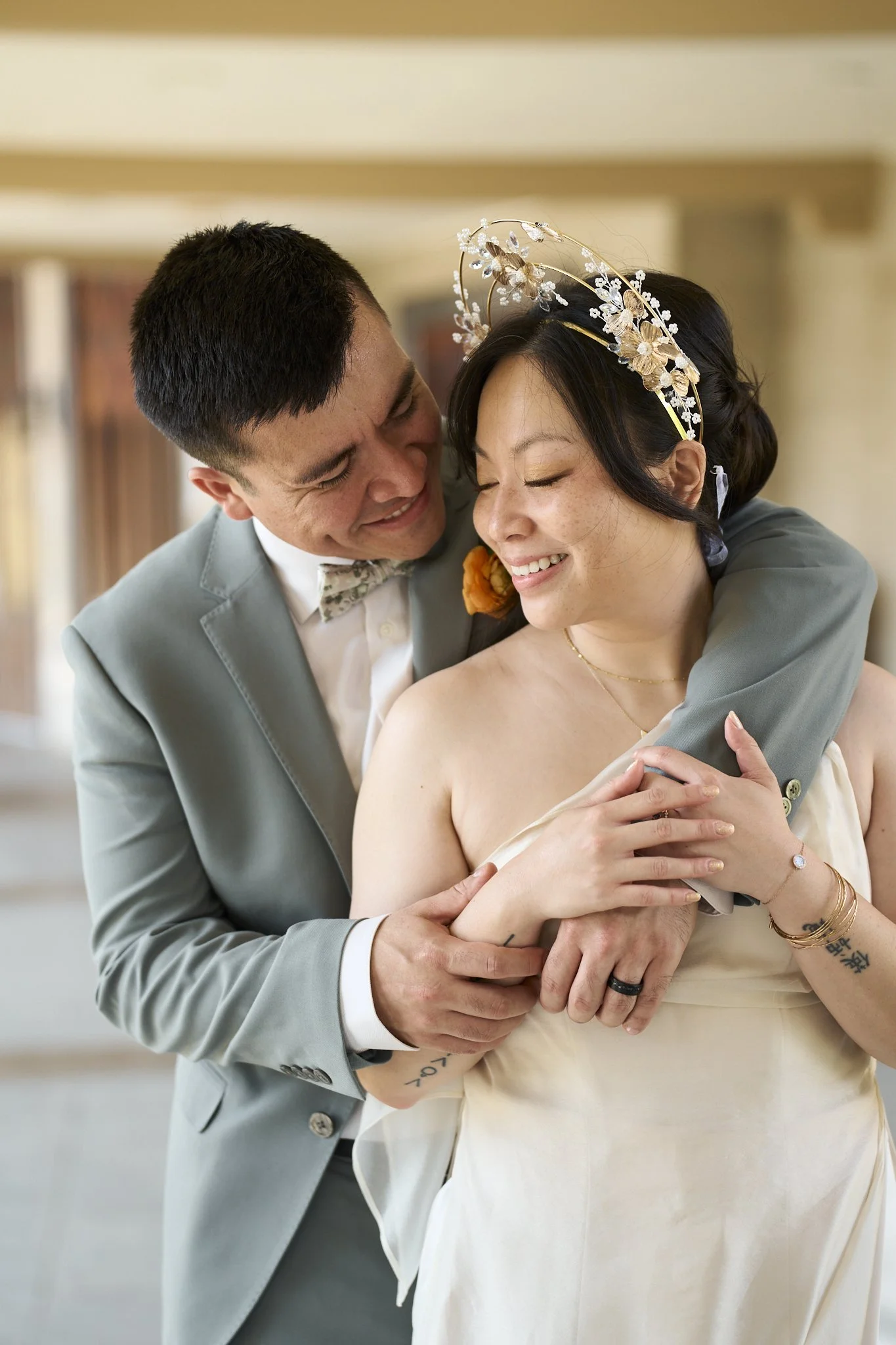 A joyful couple sharing an embrace, the man wearing a light gray suit and a floral bow tie, the woman in a strapless dress with a floral headband, both smiling with eyes closed.