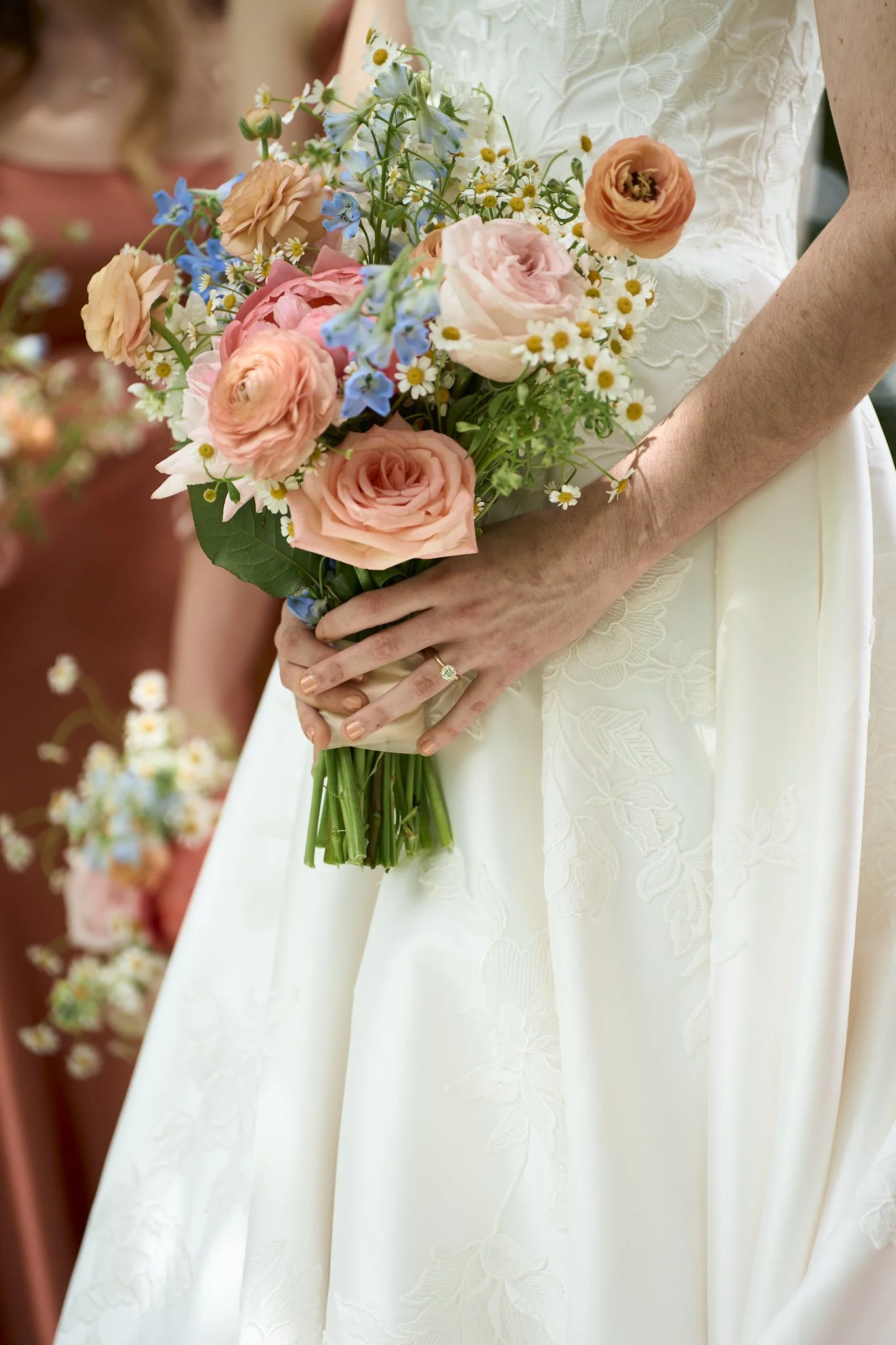 A person in a white embroidered dress holding a colorful bouquet of flowers, including pink roses, peach ranunculus, small white daisies, blue delphiniums, and greenery.