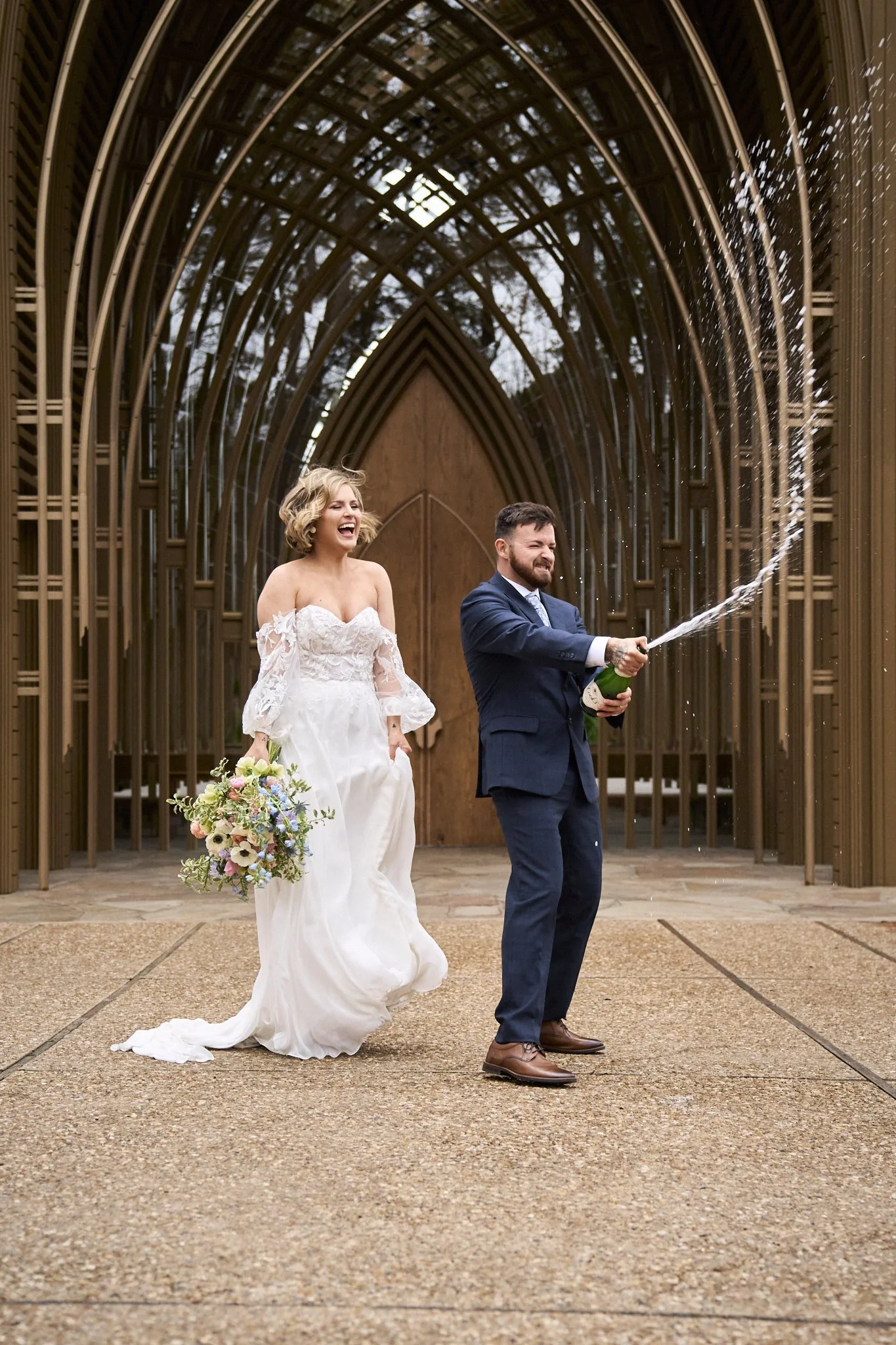 A newlywed couple celebrating outside a modern wooden chapel with arched beams. The bride wears a white lace wedding dress, holding a colorful bouquet, and is laughing joyfully. The groom, in a navy suit, is opening a champagne bottle, spraying the l