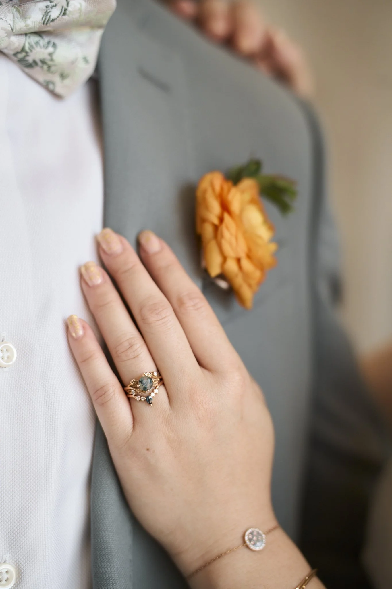Close-up of a woman's hand wearing a ring with a dark green gemstone, resting on a gray surface with a yellow-orange flower with green leaves nearby. Part of a person's white shirt and gray jacket are also visible.