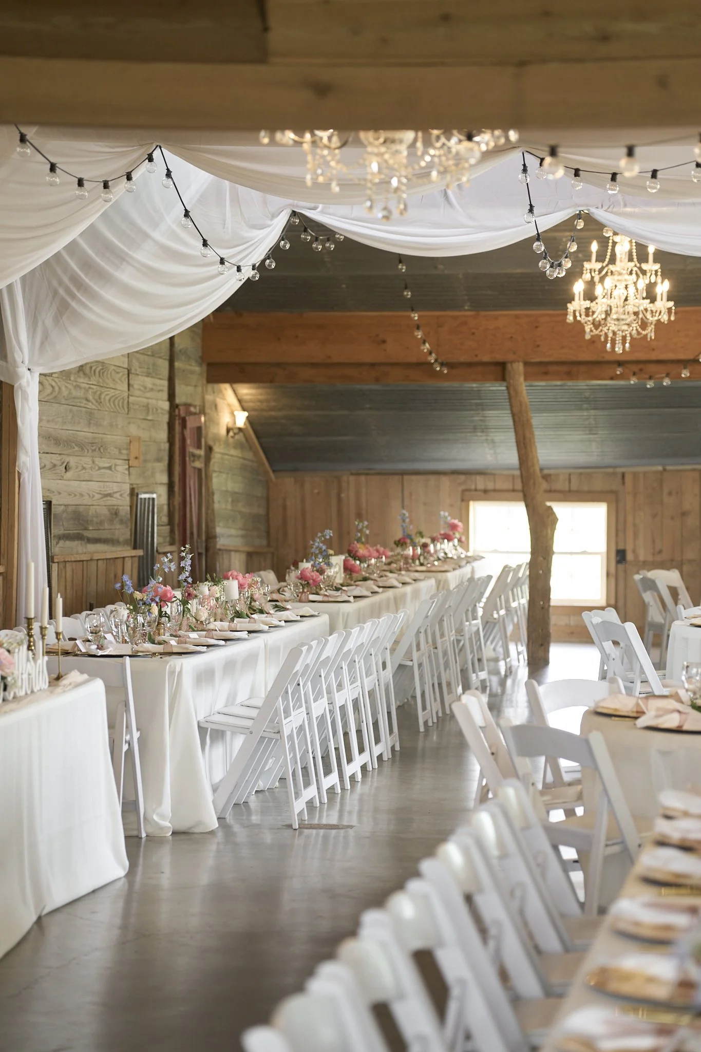 Long banquet table decorated with flowers, candles, and glassware in a rustic wooden barn with chandeliers and string lights overhead.